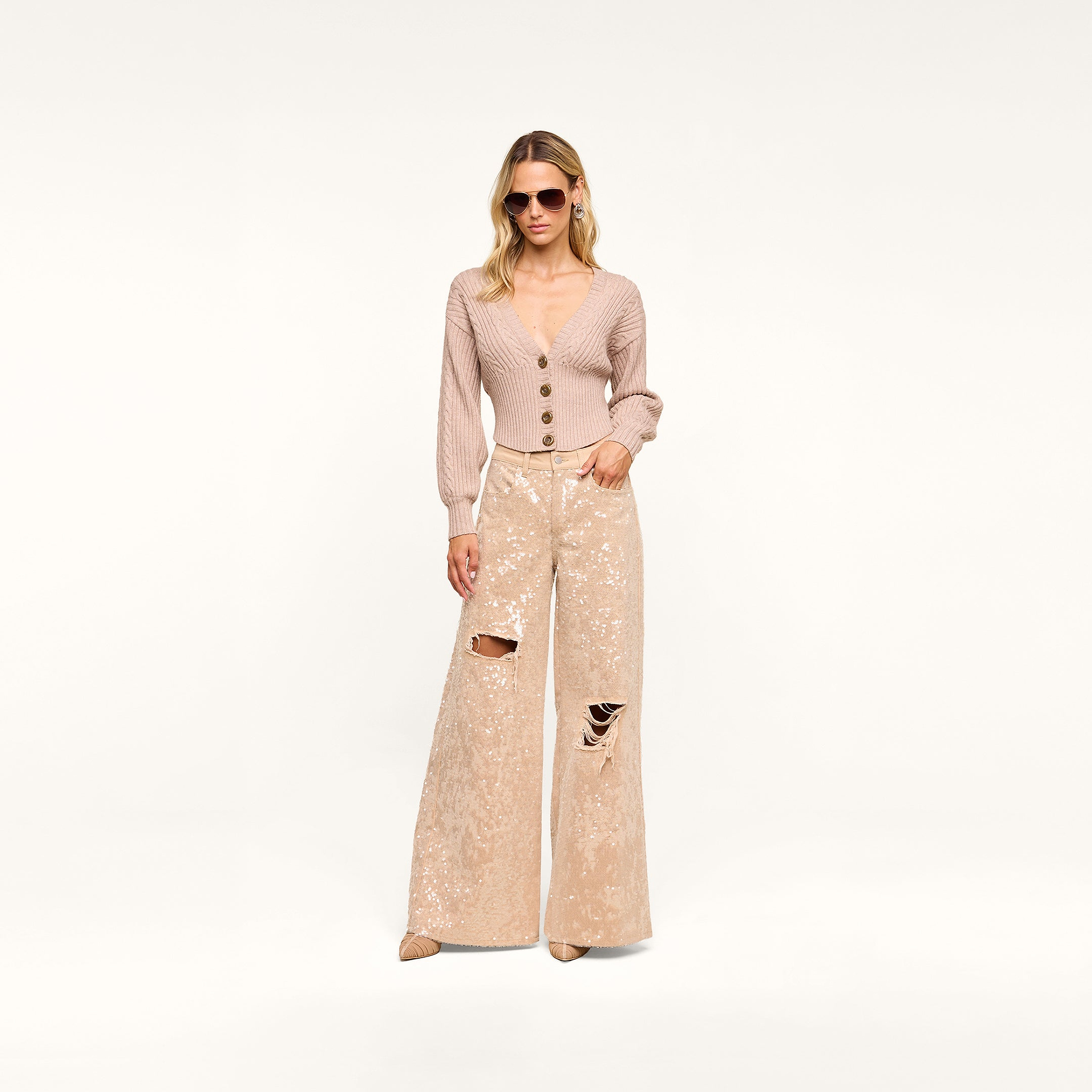 A woman wearing sunglasses, the Hail Cabled Cardigan—a beige cropped cardigan with blouson sleeves and buttons—and wide-legged, textured beige pants with ripped knees stands against a plain white background.