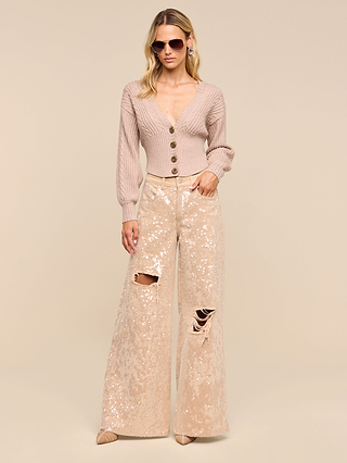 A woman wearing sunglasses and the Hail Cabled Cardigan—with large buttons and blouson sleeves—pairs it with high-waisted, wide-leg sequin pants featuring ripped knees. She stands against a plain beige background, one hand in her pocket.