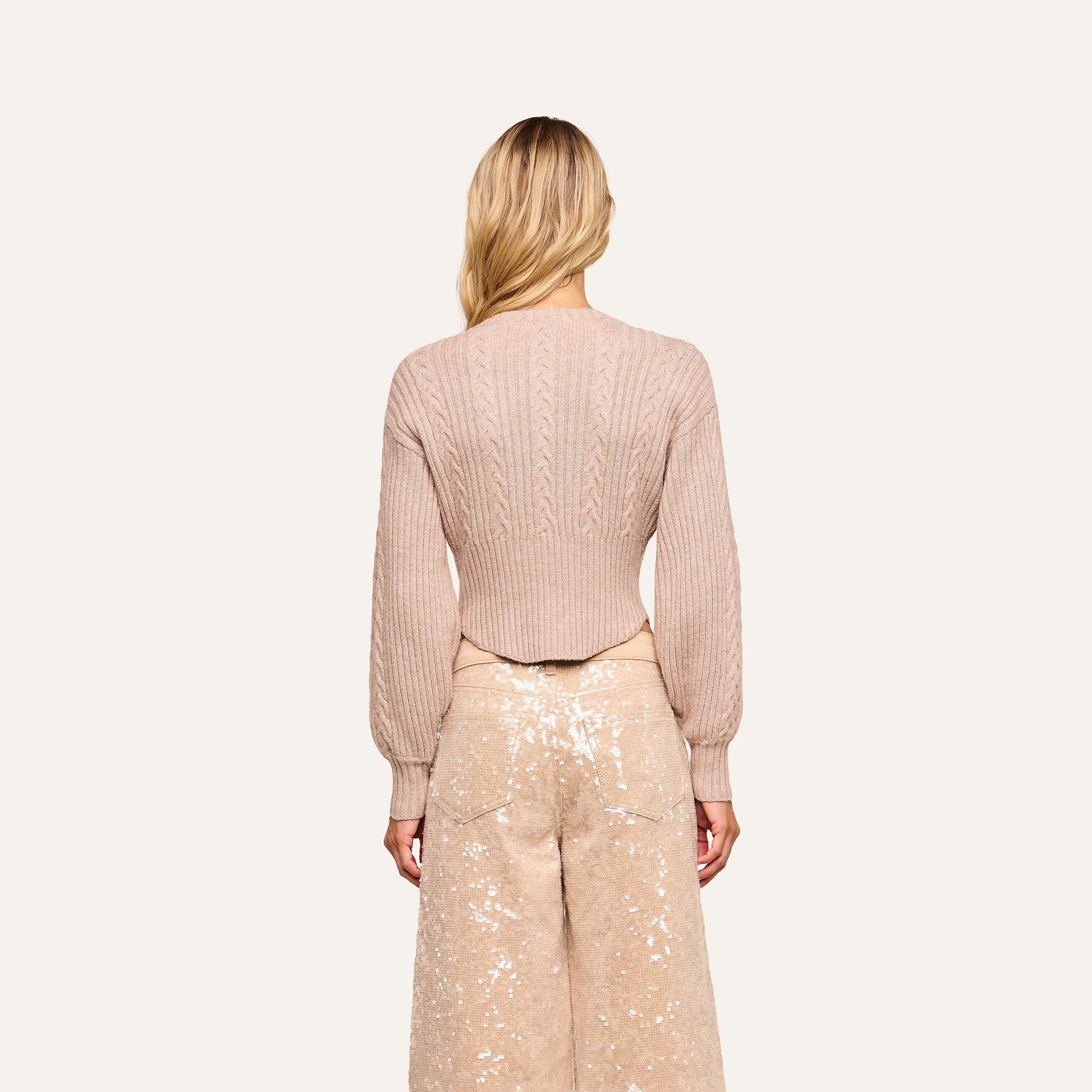 A woman with long blonde hair faces away, wearing the light pink Hail Cabled Cardigan with blouson sleeves and high-waisted, wide-leg sequined pants against a plain background.