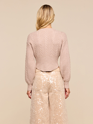 A person with long blonde hair, seen from behind, wears the Hail Cabled Cardigan in light pink—featuring blouson sleeves—paired with wide-leg, pale sequin-patterned pants, standing against a beige background.