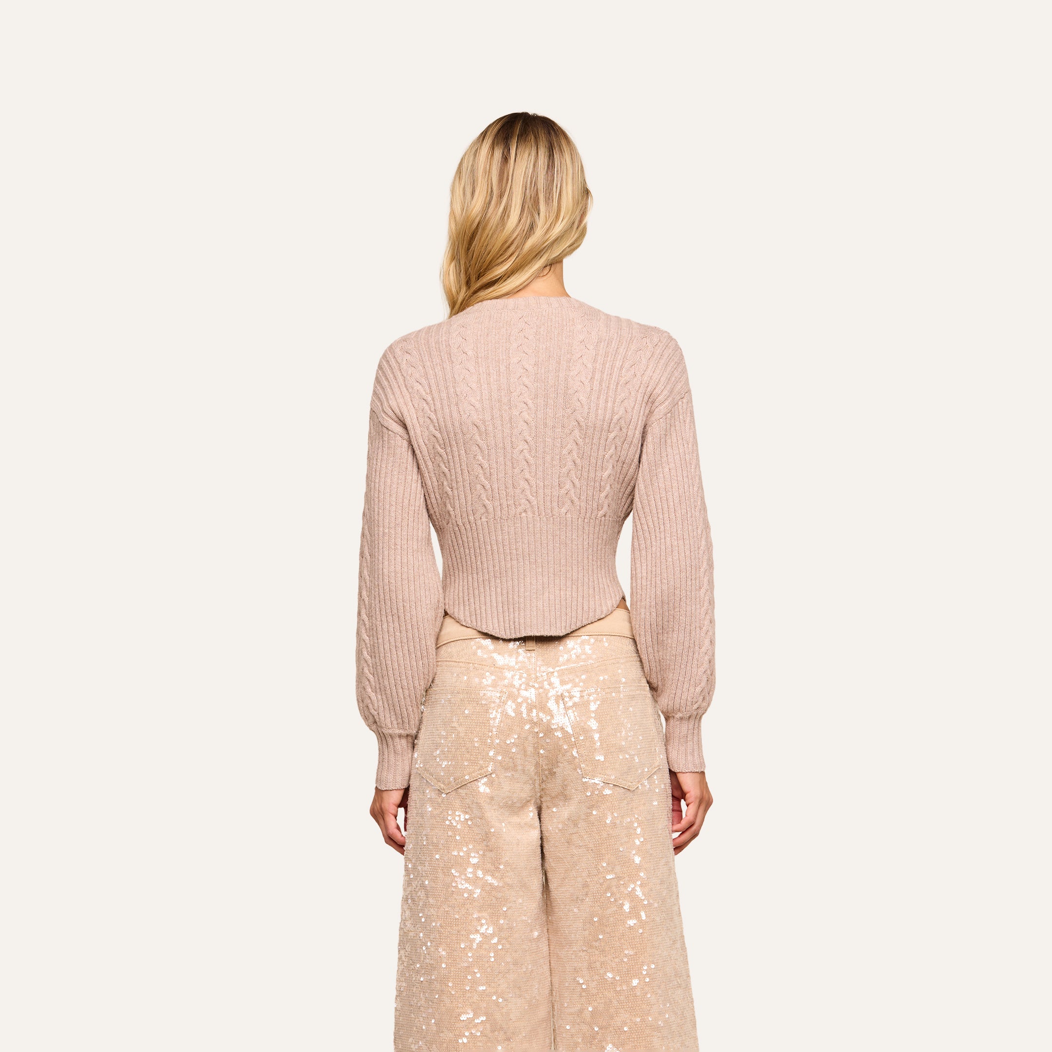 A woman with long blonde hair faces away, wearing the light pink Hail Cabled Cardigan with blouson sleeves and high-waisted, wide-leg sequined pants against a plain background.