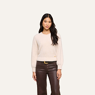 A woman stands against a plain background, wearing a relaxed fit Nimbus Pullover in light beige and brown leather pants. She has long, dark hair and a neutral expression, with her arms relaxed at her sides.