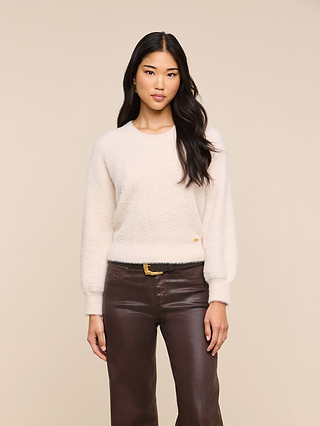 A woman with long dark hair wears the Nimbus Pullover and brown leather pants, standing against a plain beige background and looking at the camera with a neutral expression.
