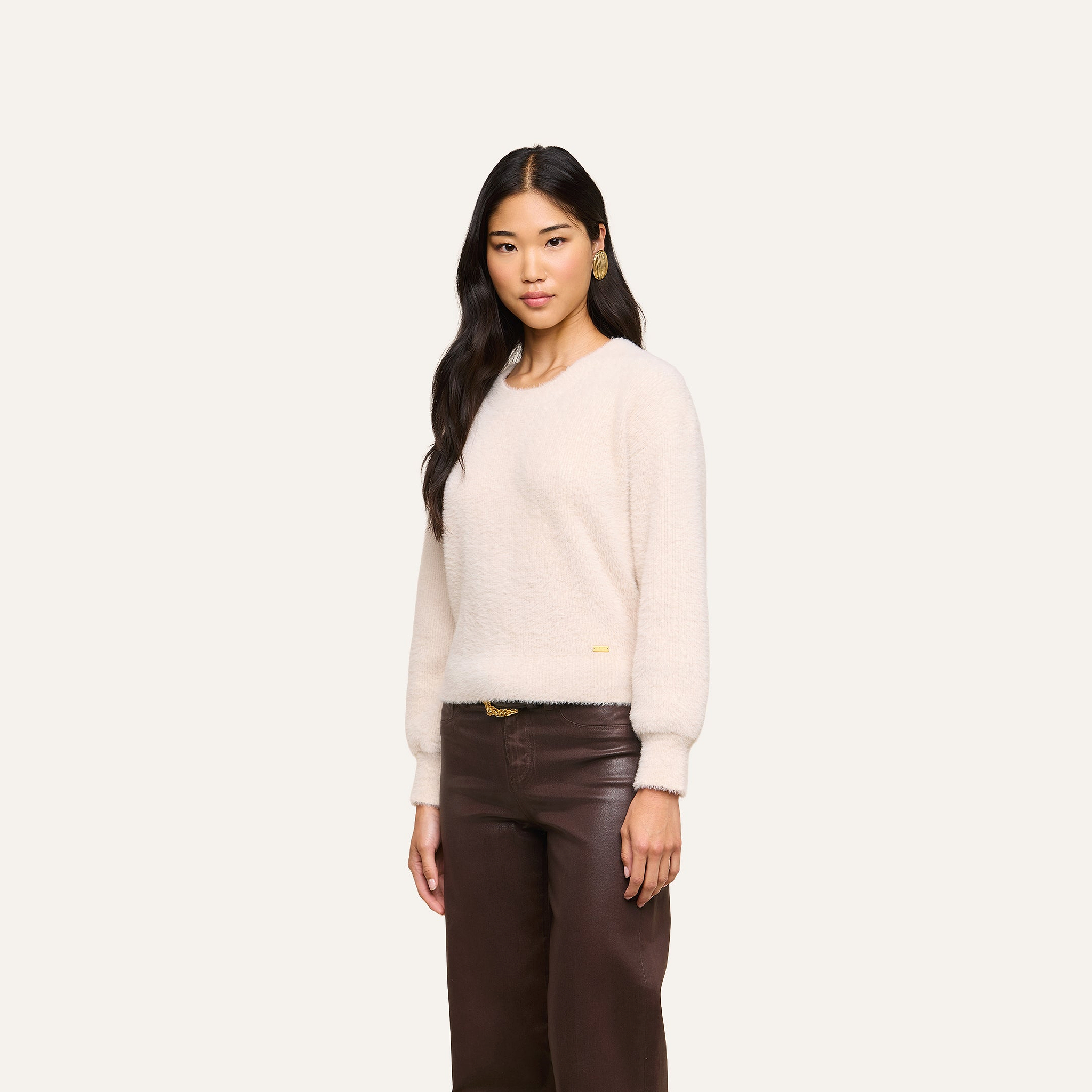 A woman with long dark hair stands against a plain light background, looking at the camera with a neutral expression, wearing the cloud-soft Nimbus Pullover sweater and brown leather pants.