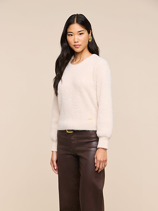 A woman with long dark hair wears the Nimbus Pullover—a relaxed fit, cloud-soft sweater in cream—paired with brown leather pants. She stands against a plain beige background, looking at the camera.