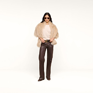 A woman stands confidently against a plain white background, wearing sunglasses, brown leather pants, a light top, and the Nimbus Pullover—a beige faux fur jacket with a relaxed fit.