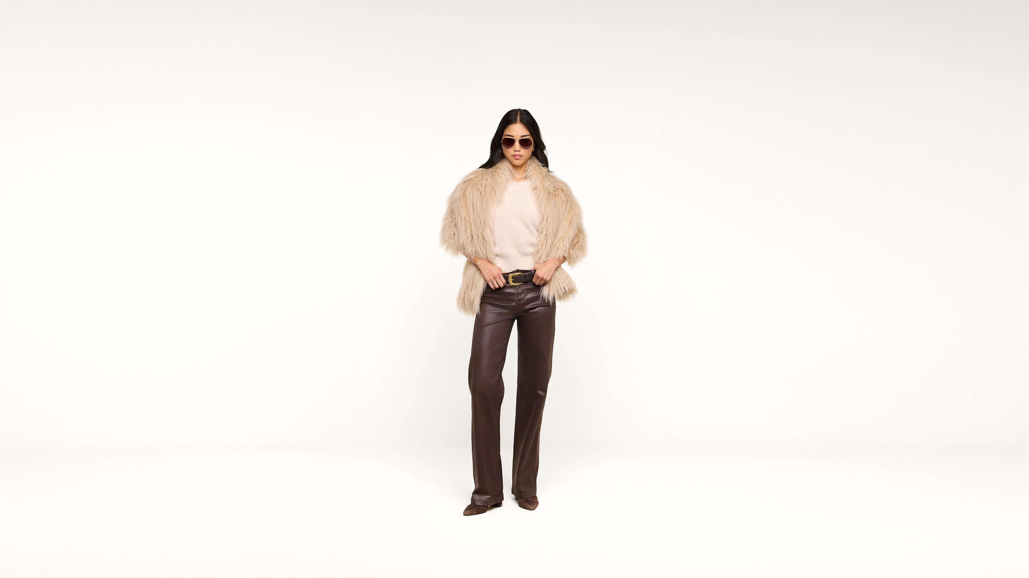 A woman wearing sunglasses, a beige fur jacket, light top, and dark brown leather pants stands against a plain white background, posing with hands in her pockets.