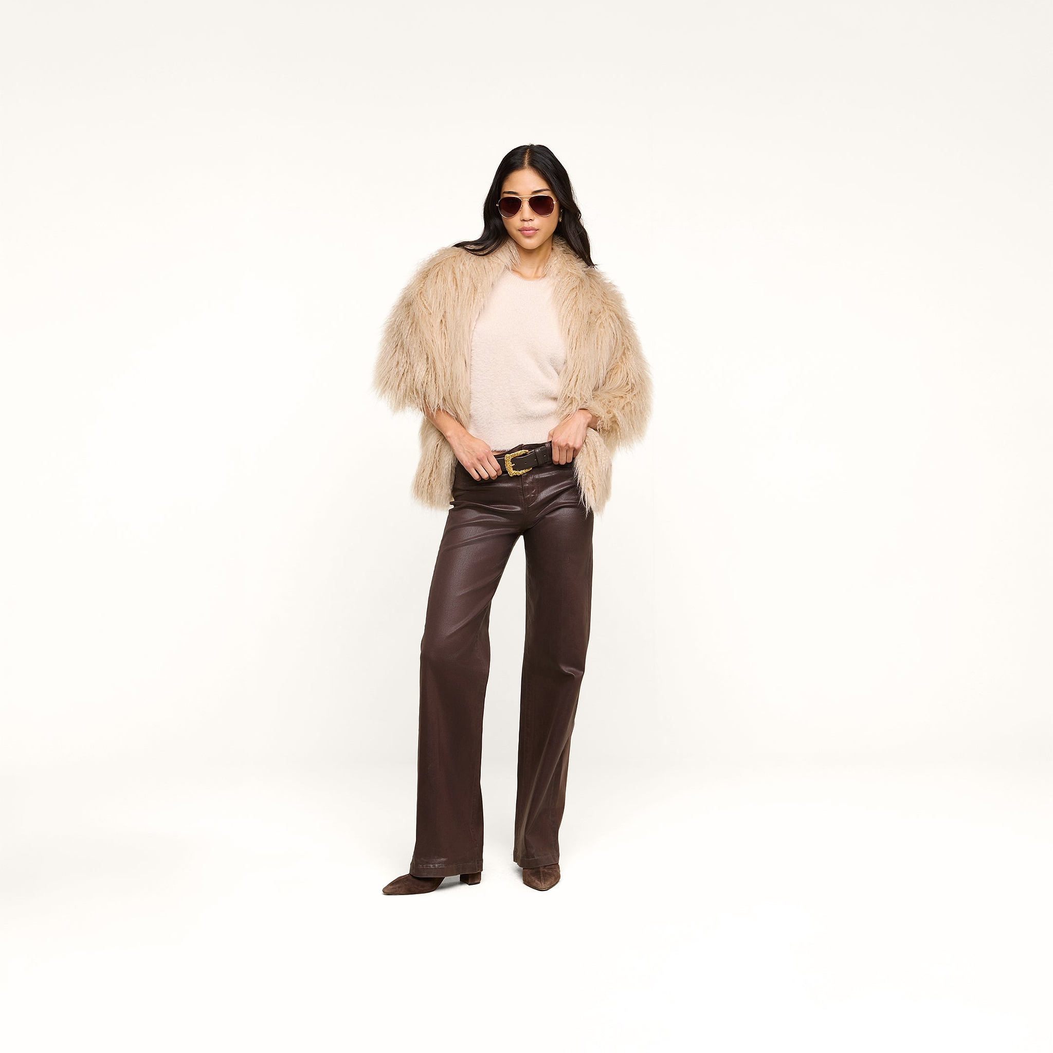 A woman wearing a beige faux fur jacket, brown leather pants, sunglasses, and the Nimbus Pullover—an ultra-soft cream cloud-soft sweater—stands confidently against a plain white background.