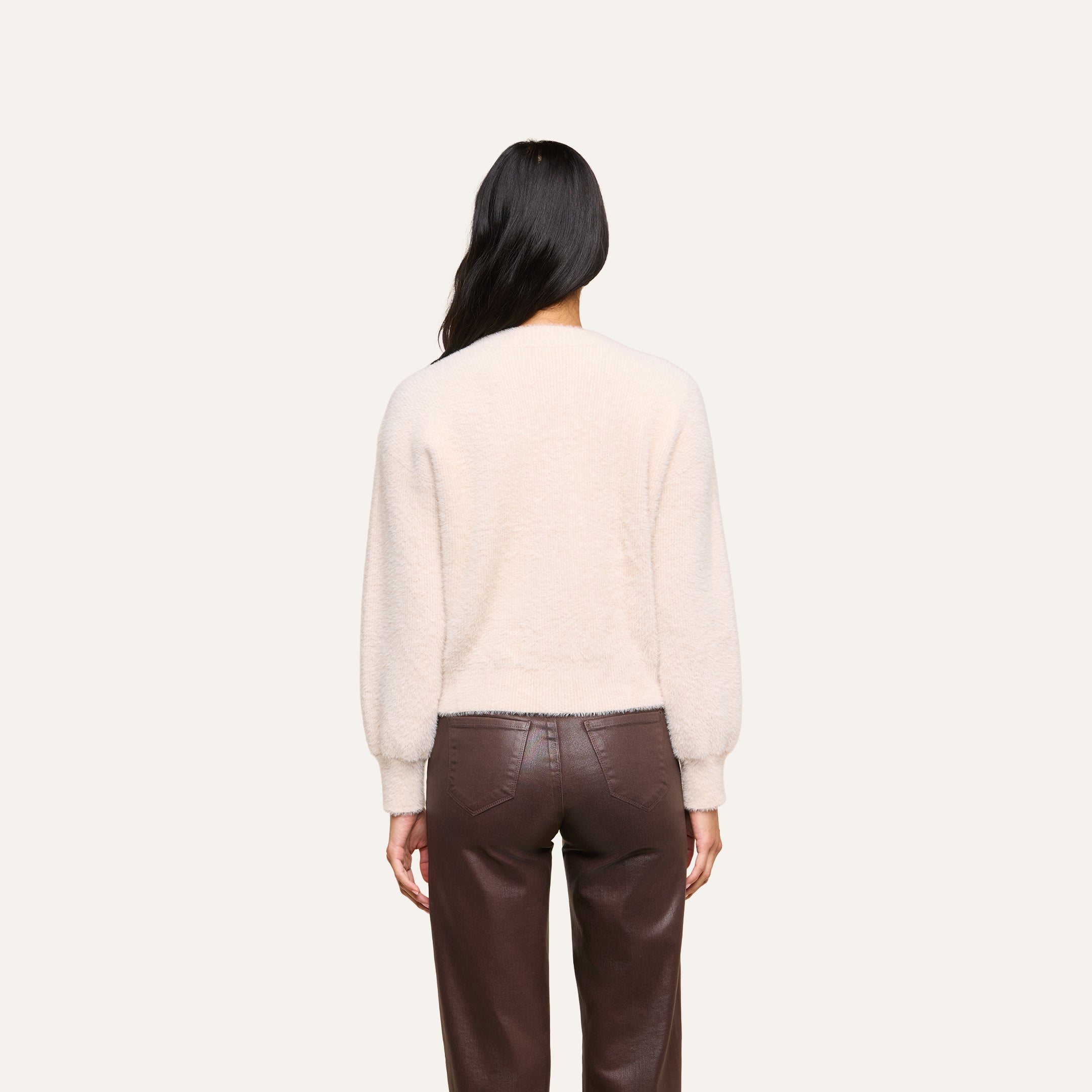 A person with long dark hair is shown from the back, wearing the Nimbus Pullover, a relaxed fit cream-colored fuzzy sweater with balloon sleeves, paired with high-waisted brown leather pants and standing against a plain light background.