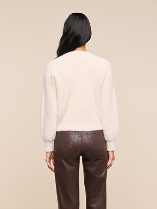 A person with long dark hair, seen from behind, is wearing the Nimbus Pullover in a cream color along with brown leather pants, standing against a plain beige background.