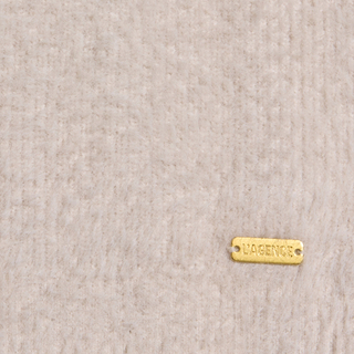 A small gold metal tag engraved with LAGENCE is attached to a soft, light beige textured fabric in the bottom right corner, adding a signature touch to the Nimbus Pullover.