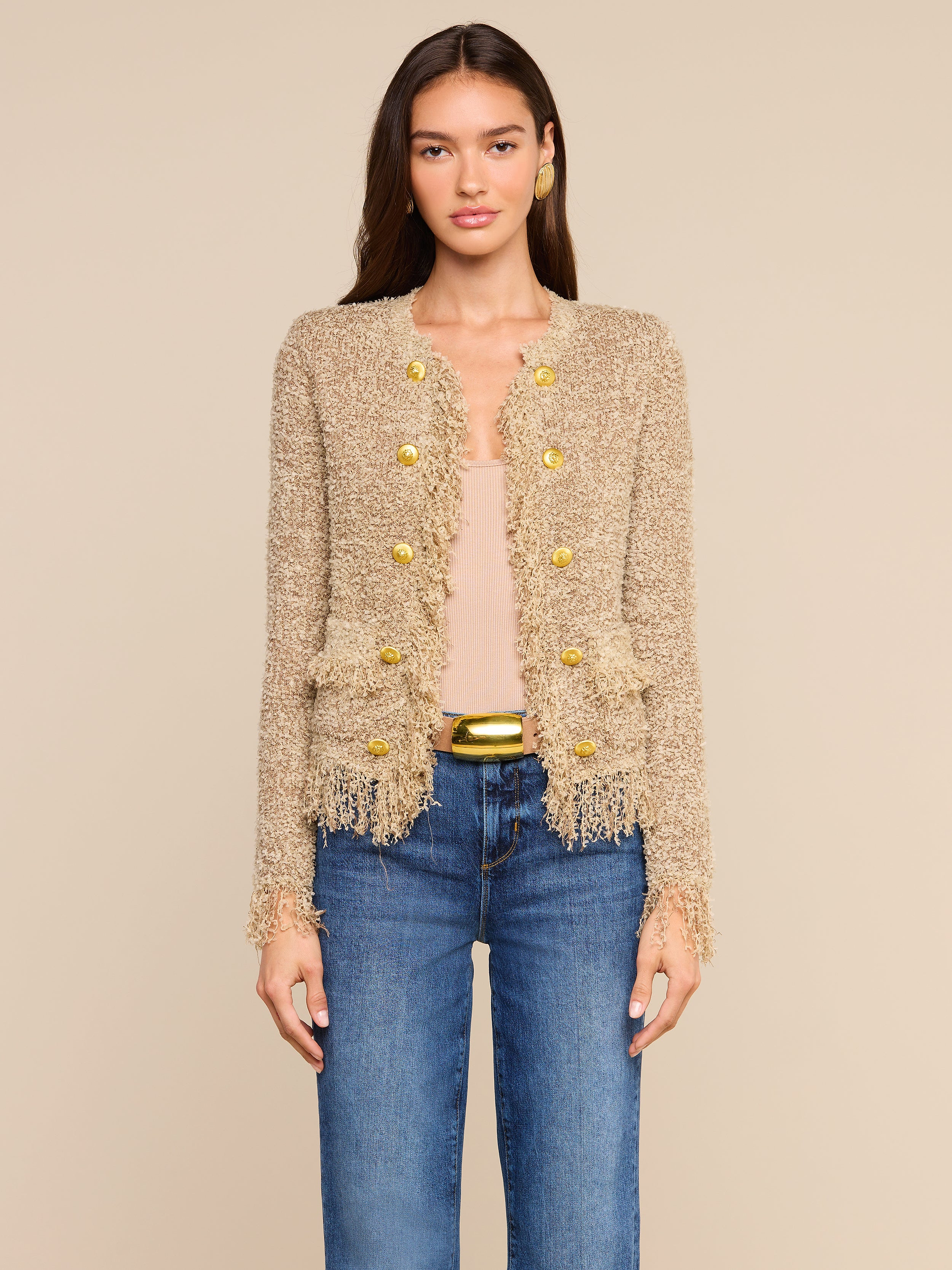 A woman wearing the Mireille Fringe Cardigan, which features textured beige fabric with metallic threads and gold buttons, paired with a light-colored top and blue jeans, stands against a plain light background.