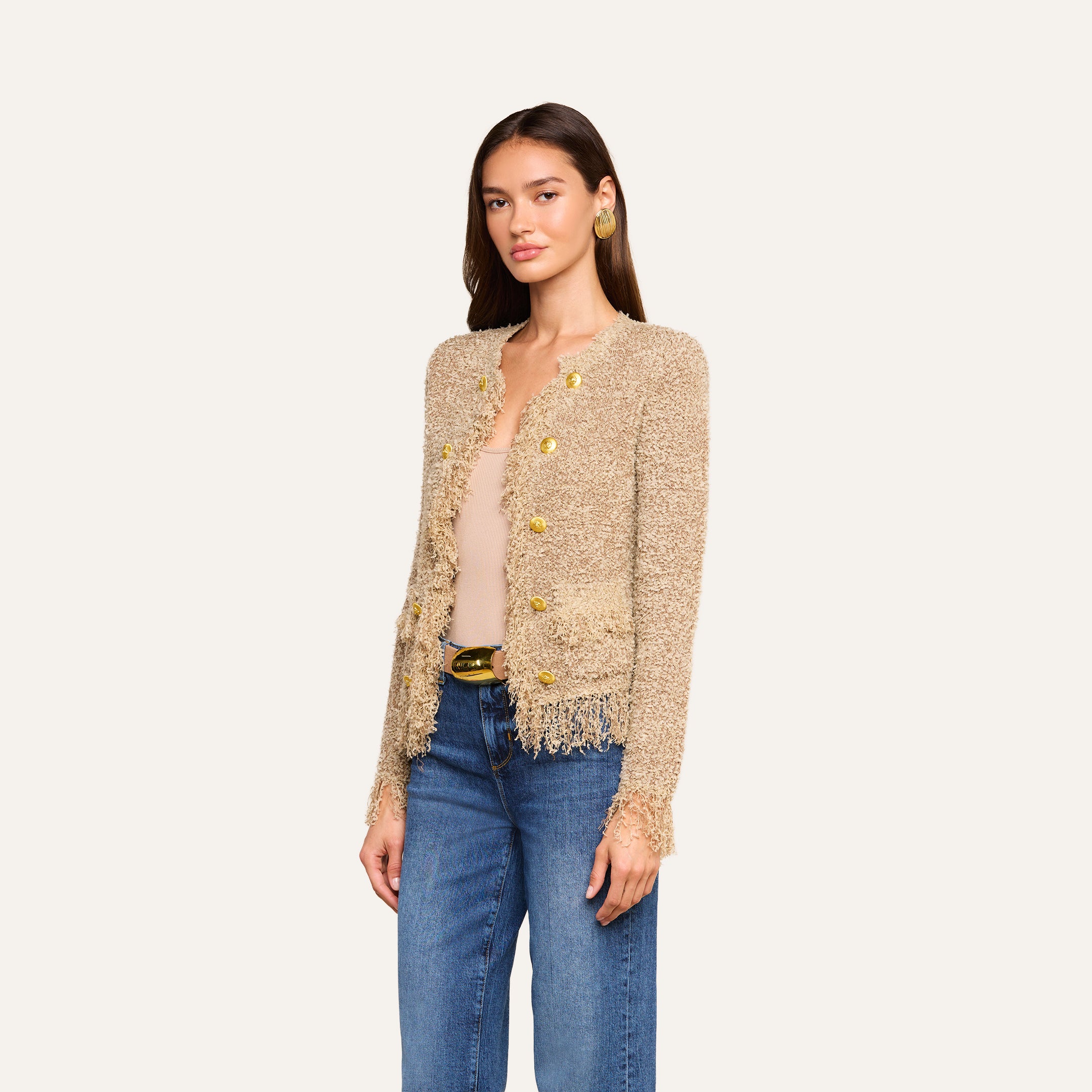 Woman wearing the Mireille Fringe Cardigan, featuring a textured beige confetti knit with subtle metallic threads and gold buttons, layered over a nude top and blue jeans, standing against a plain light background.