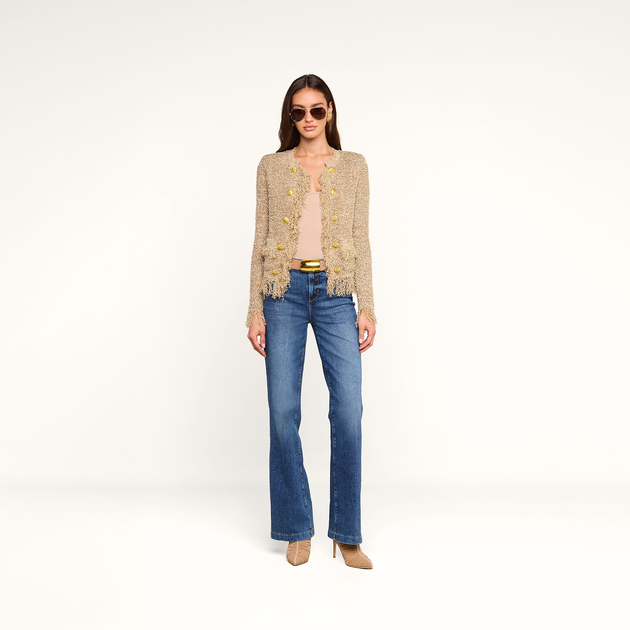 A woman wearing sunglasses, a Mireille Fringe Cardigan with flower detailing, a light top, blue jeans, and tan heels stands against a plain white background.