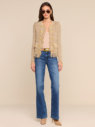 Woman wearing sunglasses, a beige Mireille Fringe Cardigan with gold buttons and subtle metallic threads, a light top, blue jeans, a gold-buckled belt, and beige heels, standing against a plain light background.