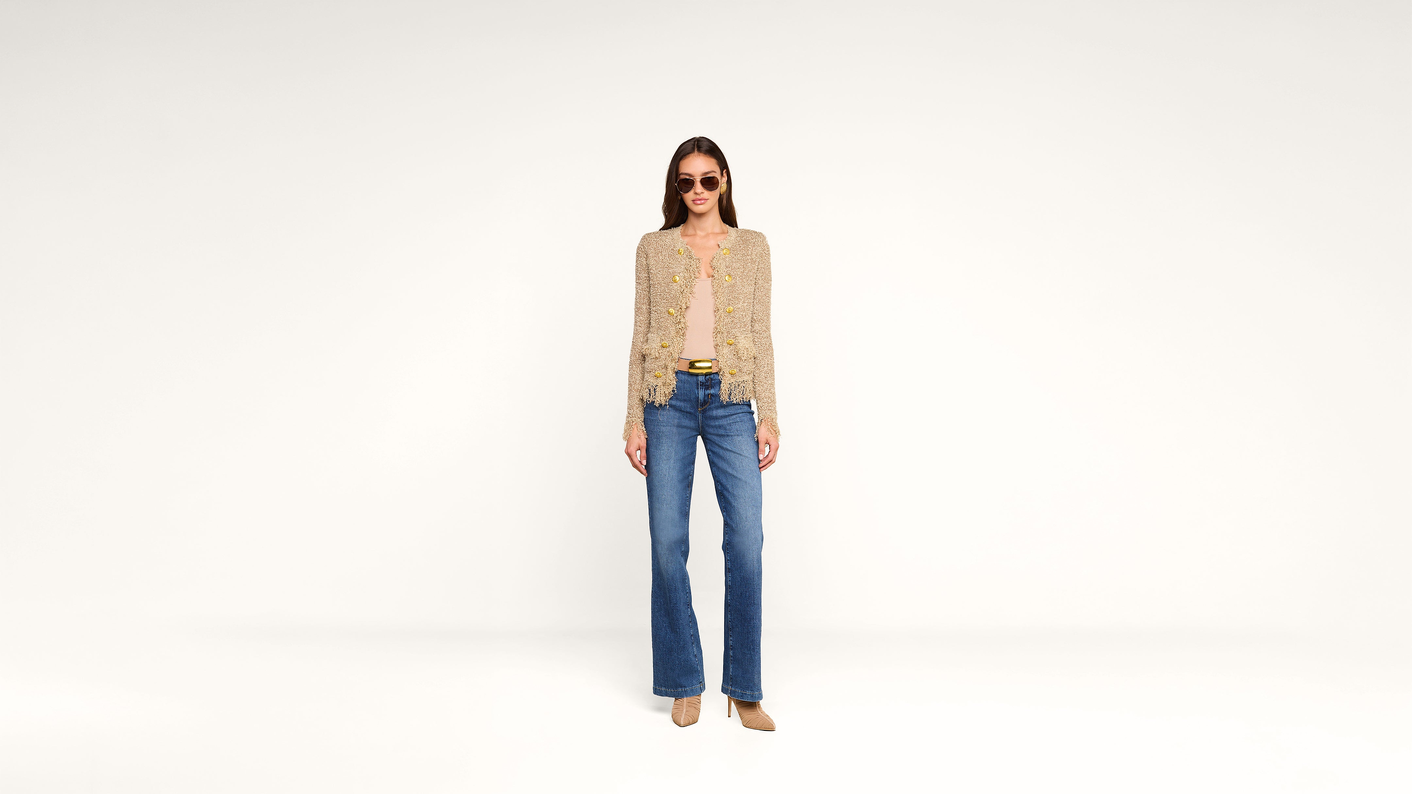 A woman wearing sunglasses, a tan cardigan, a yellow top, blue jeans, and beige heels stands facing forward against a plain white background.