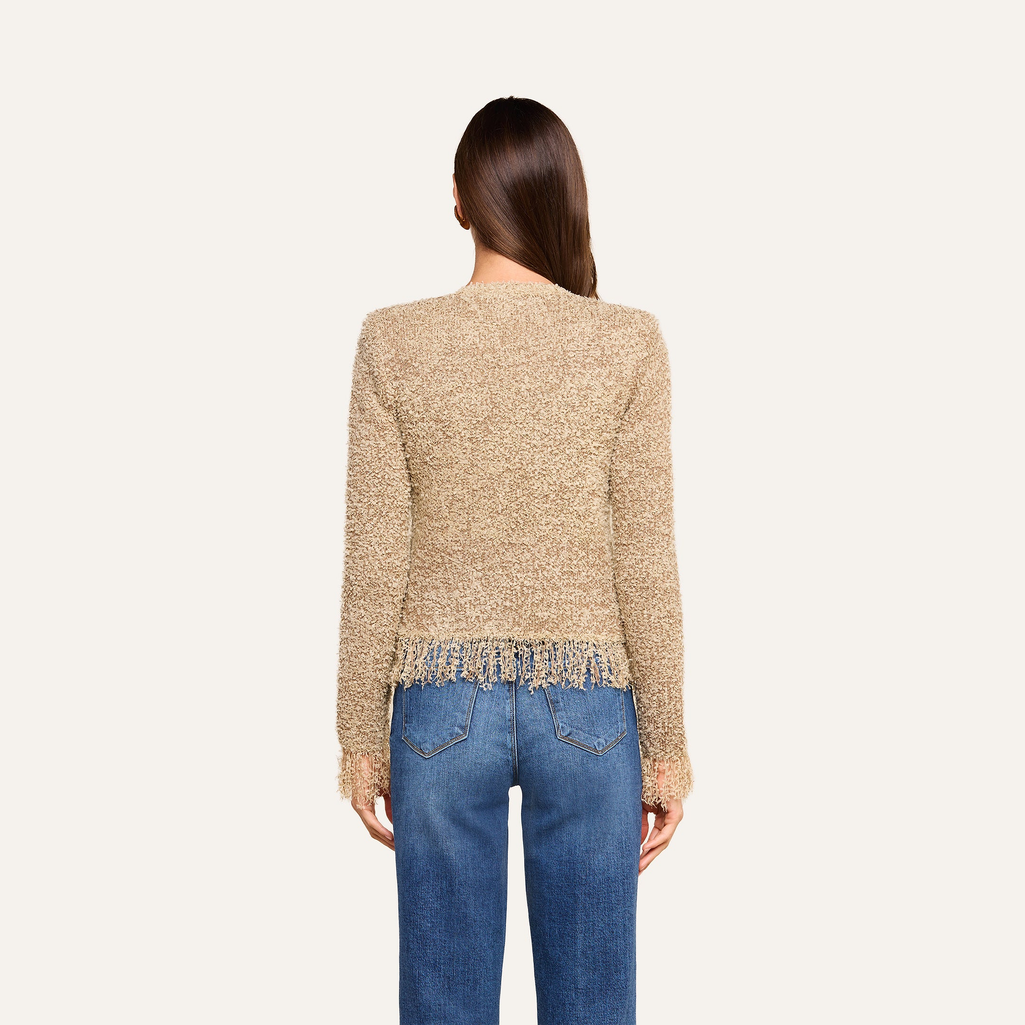 A woman with long brown hair, seen from behind, wears the Mireille Fringe Cardigan—a confetti knit tan sweater with fringe at the hem—paired with blue jeans. The background is off-white.