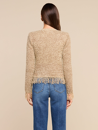 A woman with long brown hair, seen from behind, wears the Mireille Fringe Cardigan—a textured beige piece featuring fringe at the hem and cuffs and subtle metallic threads—paired with blue jeans, standing against a plain beige background.