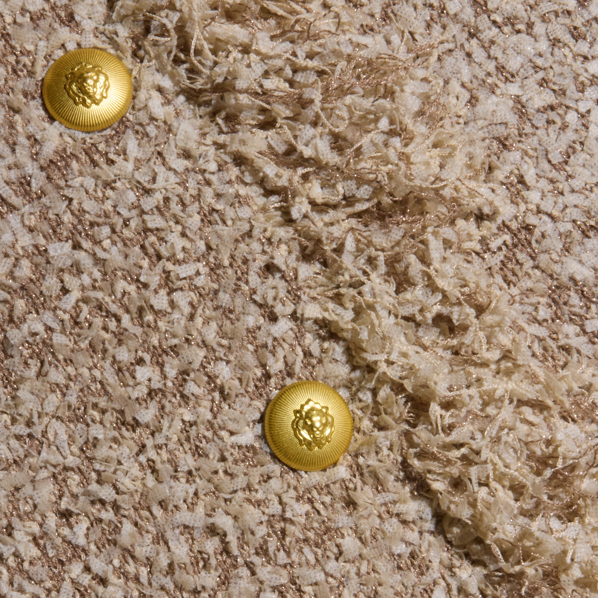 Close-up of the Mireille Fringe Cardigan’s textured beige confetti knit fabric, featuring two shiny gold lion’s head buttons and diagonal sections of smooth and fringed detailing.