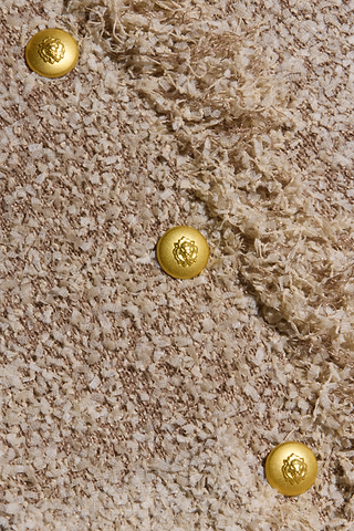 Close-up of the textured beige confetti knit fabric from the Mireille Fringe Cardigan, featuring three shiny gold buttons arranged diagonally. Each button is embossed with a raised emblem design at the center, adding an elegant touch characteristic of this style.