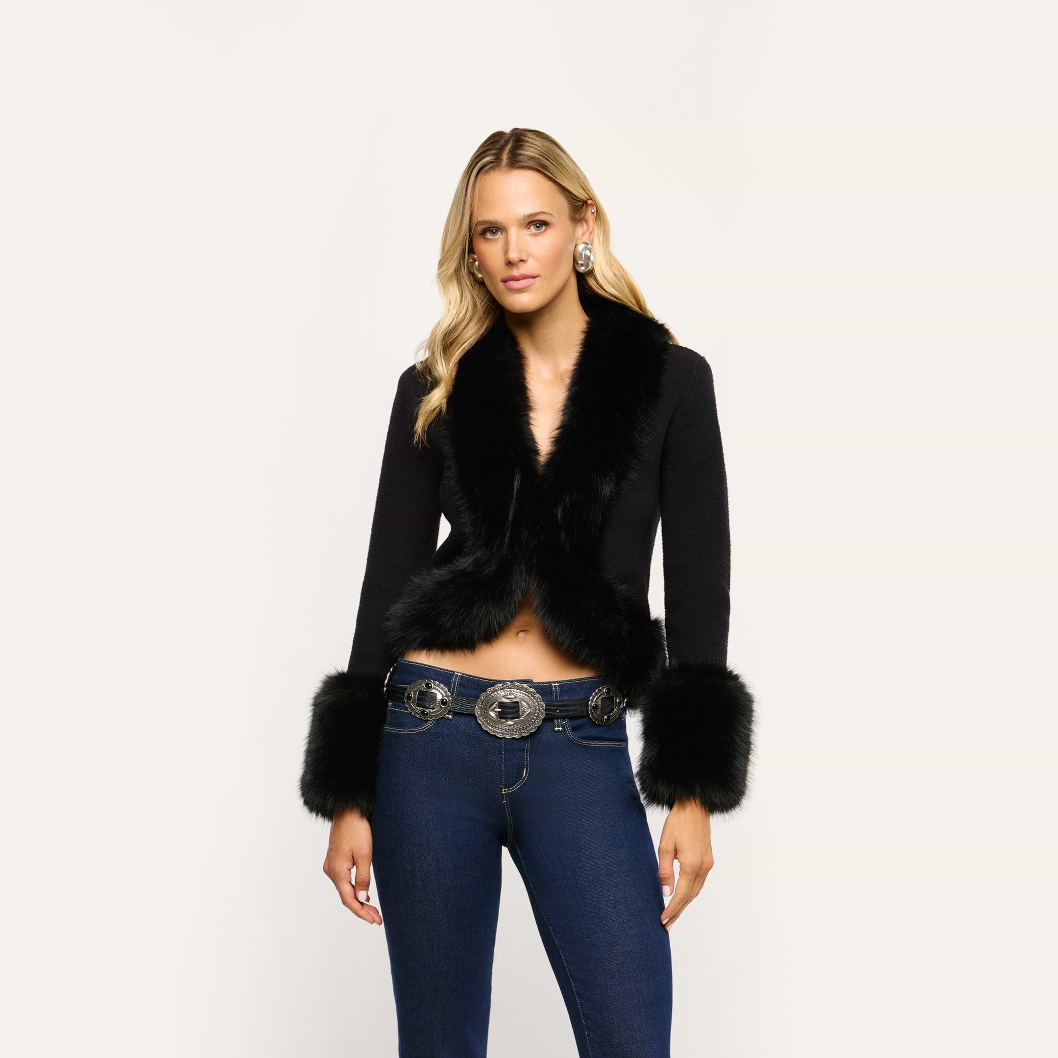 The Irma Cardigan, a cropped black Y2K style with faux fur trim on the collar and cuffs, worn over a denim belt with a large metal buckle and dark blue jeans.