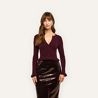 A woman wears a burgundy ribbed, cropped Finian Cardigan Top with a notched neckline and ruffled cuffs, paired with a dark sequined midi skirt.