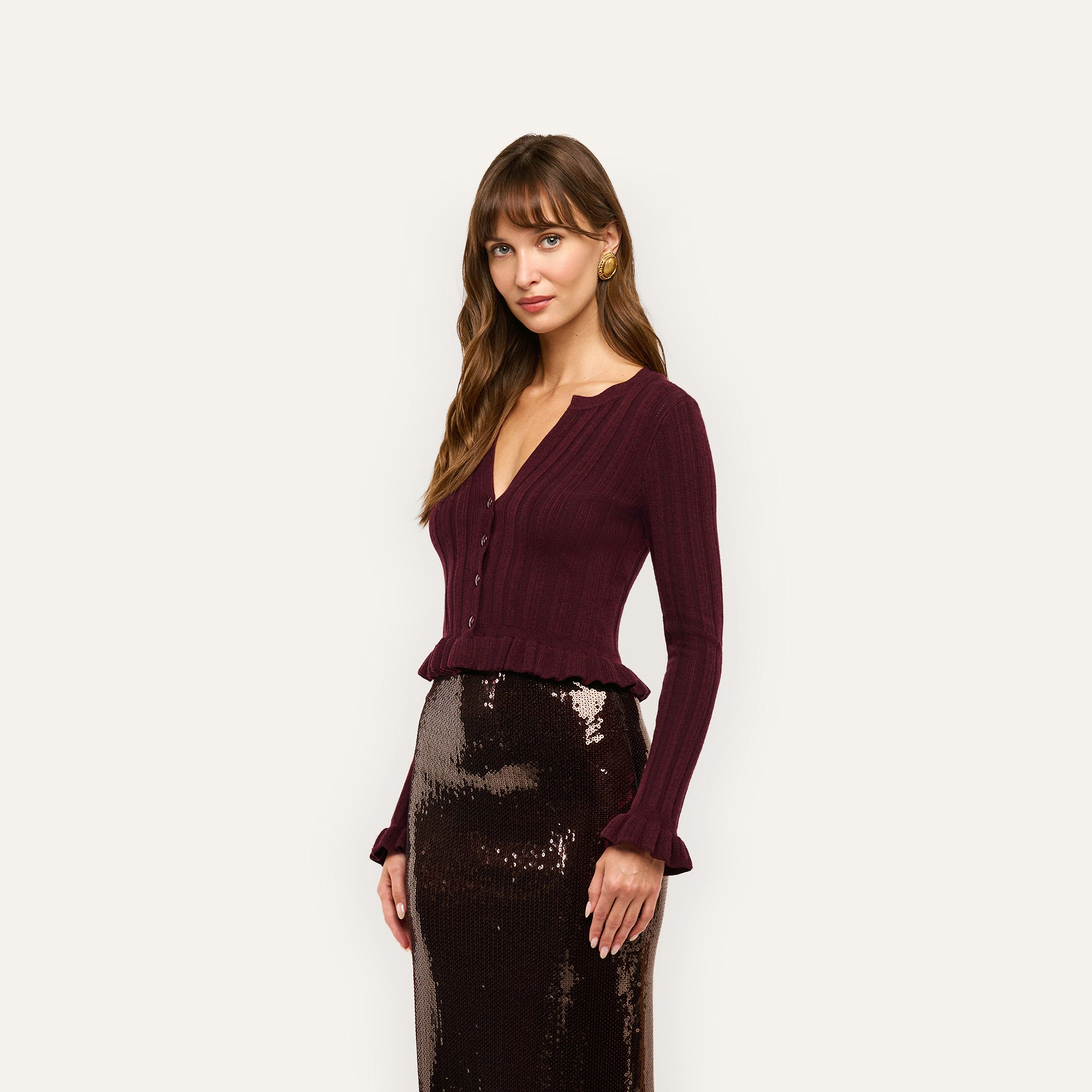 A woman wearing a dark maroon, lightweight, cropped ribbed-knit Finian Cardigan Top with long sleeves, a V-neck, and ruffled hems, paired with a shimmering dark brown sequin skirt.