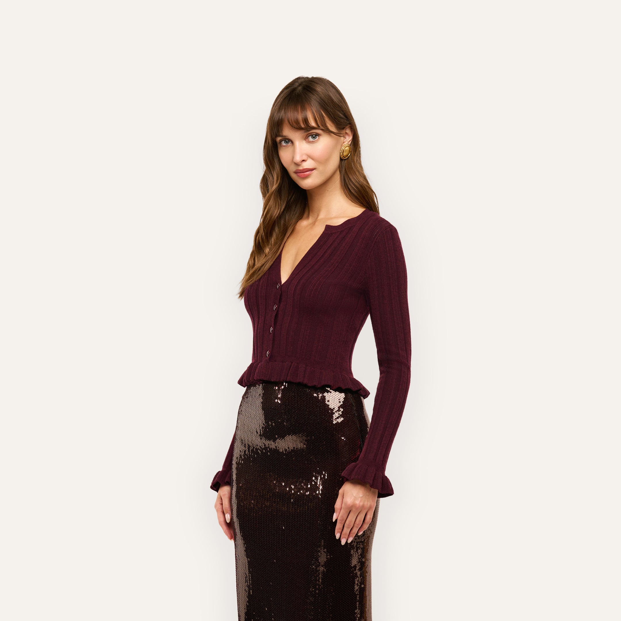 A woman wearing a dark maroon, lightweight, cropped ribbed-knit Finian Cardigan Top with long sleeves, a V-neck, and ruffled hems, paired with a shimmering dark brown sequin skirt.