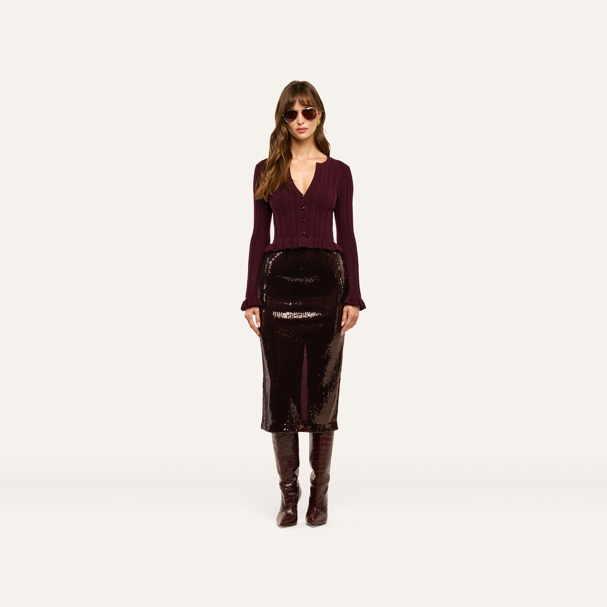 A woman wears a lightweight, ribbed burgundy Finian Cardigan Top with a ruffled hem, a dark sequined midi skirt, and knee-high boots.
