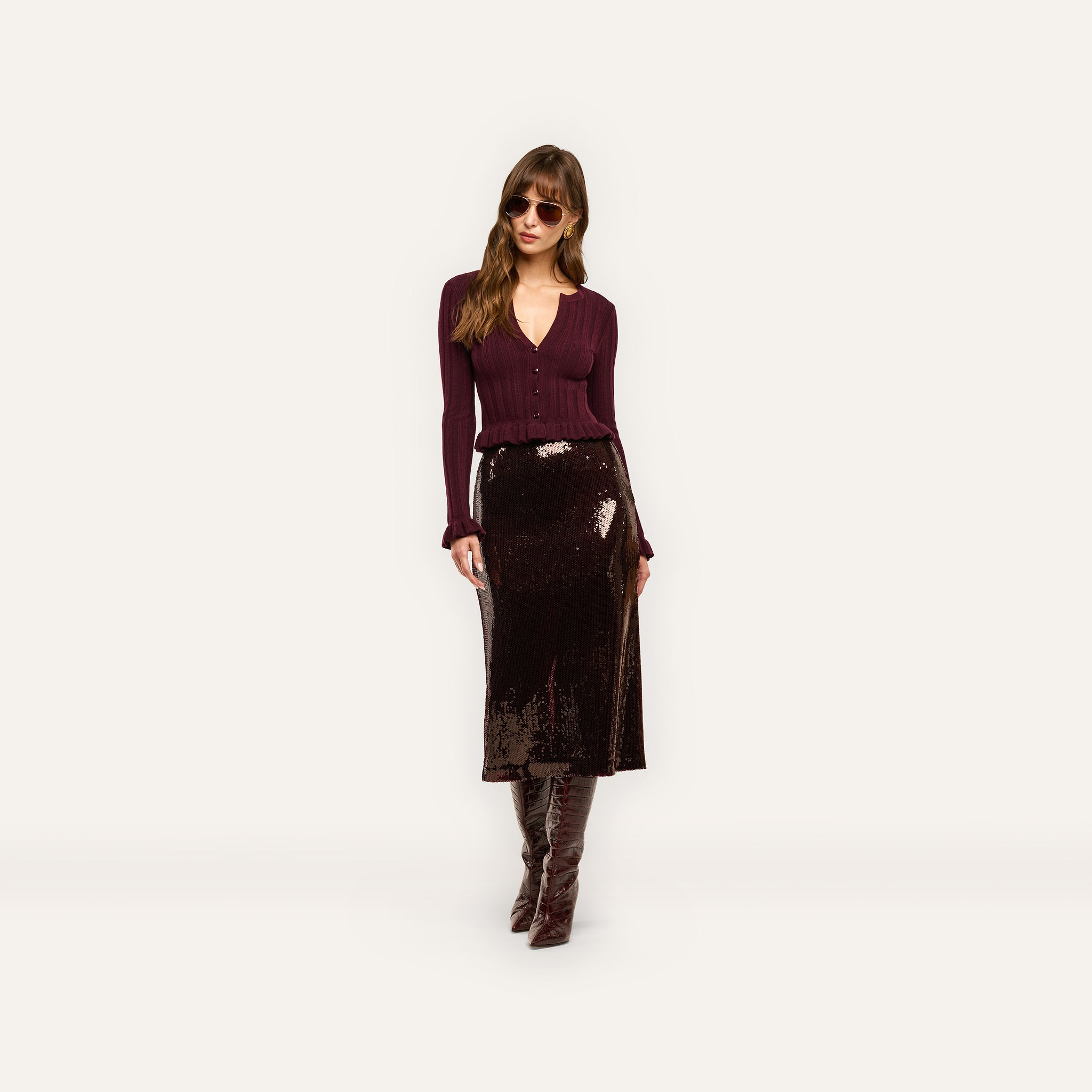 A woman wearing a deep red Finian Cardigan Top, a dark sequined midi skirt, and knee-high boots.