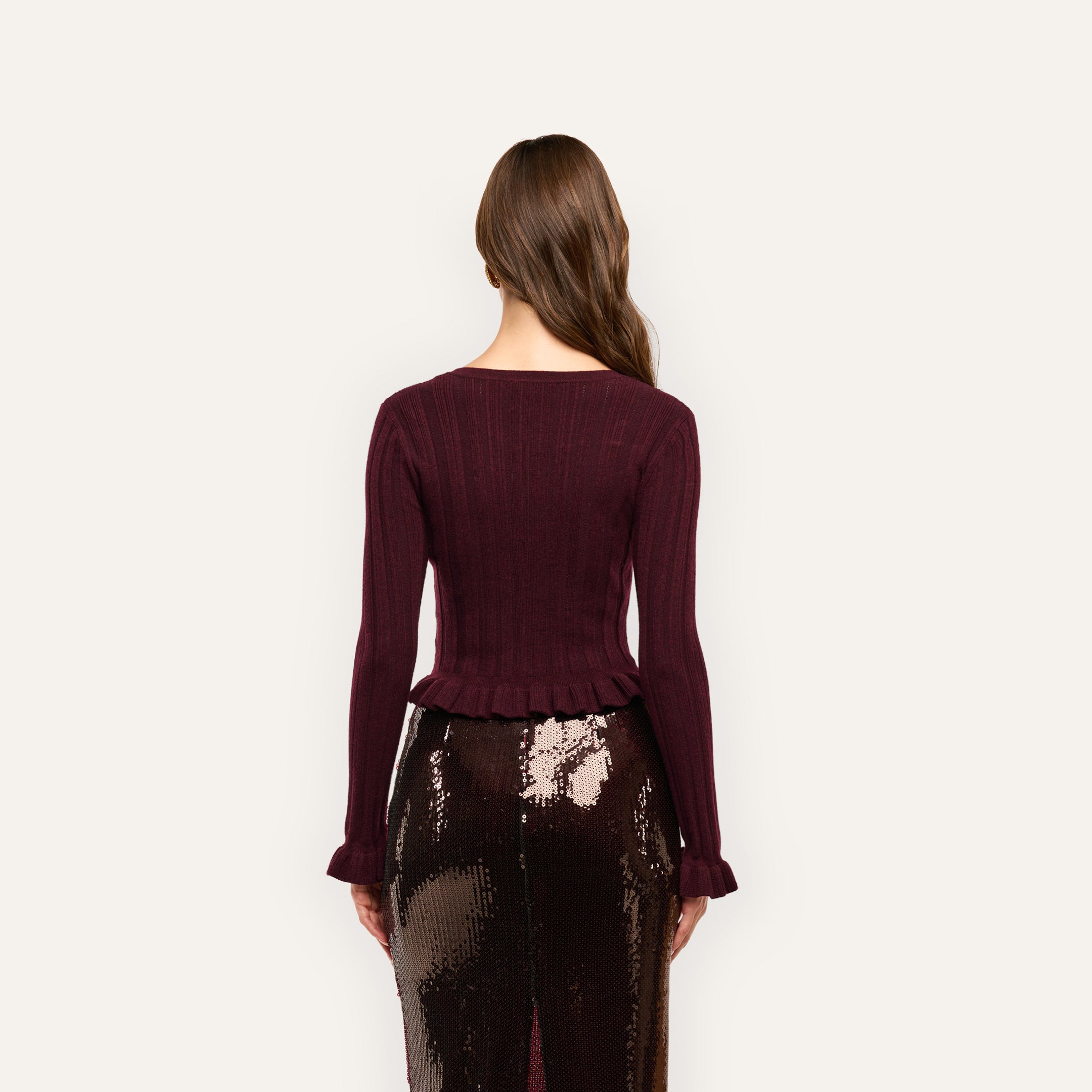 Back view of a person wearing a Finian Cardigan Top, a lightweight, ribbed burgundy cardigan with a cropped, ruffled hem, paired with a shiny, dark-toned sequined skirt.