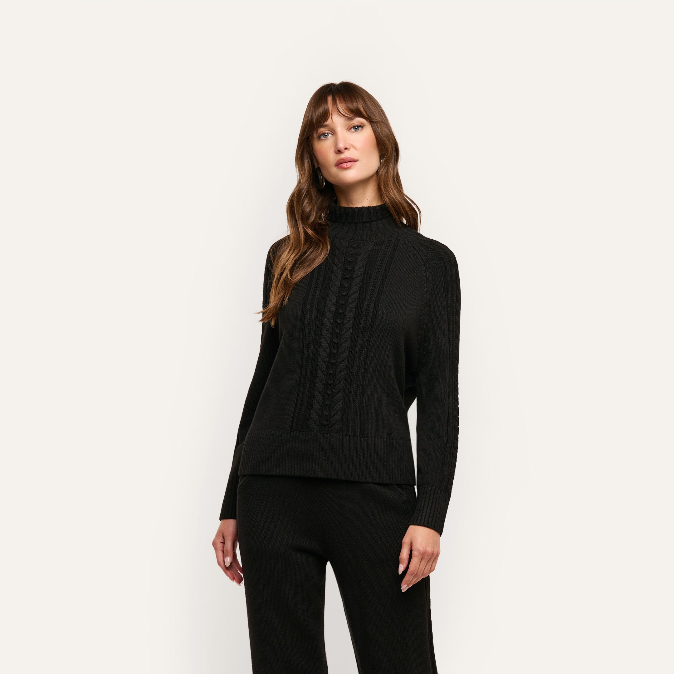 A person wearing the Sandrina Turtleneck, a relaxed-fit, black long-sleeved sweater featuring a central cable-knit pattern.