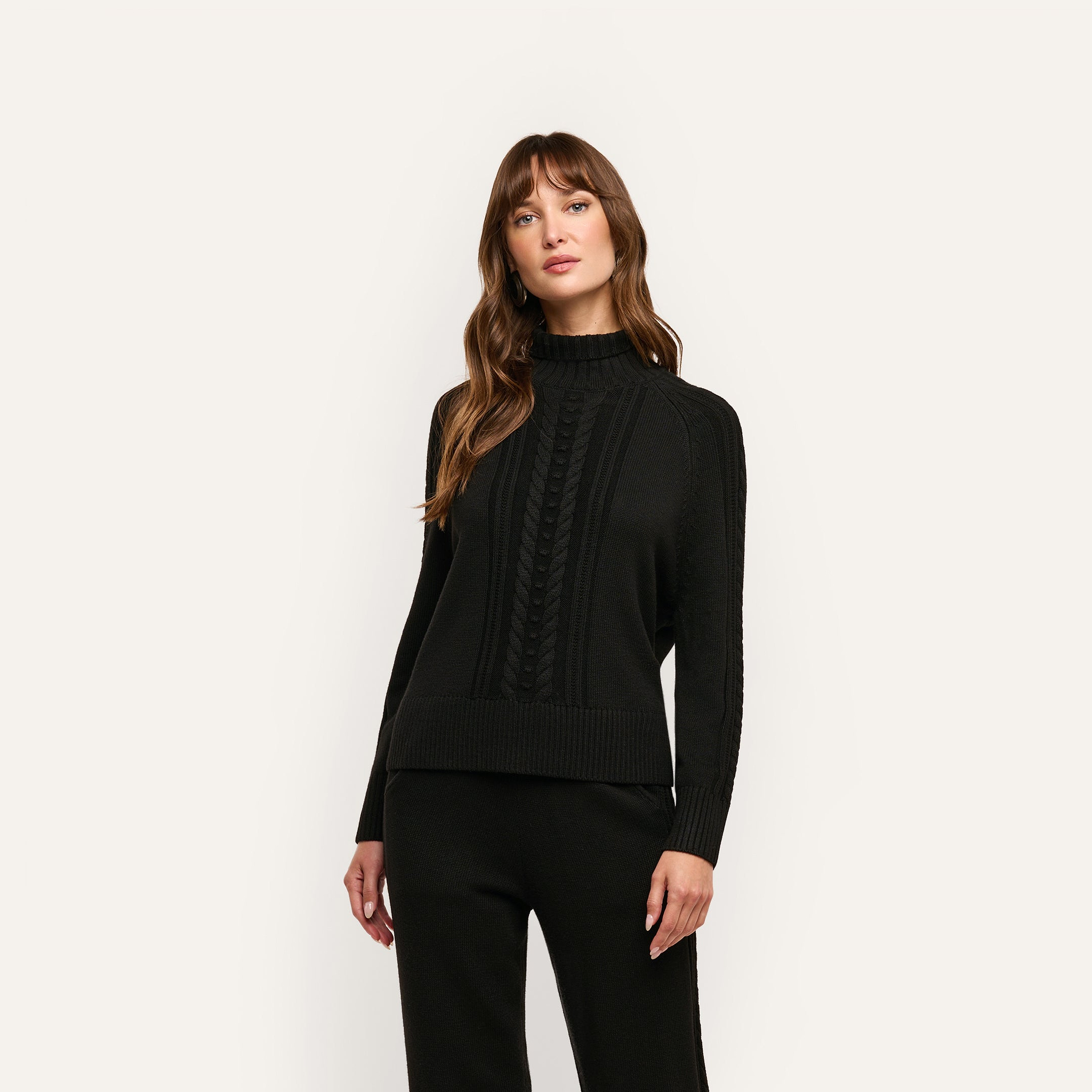 A person wearing the Sandrina Turtleneck, a relaxed-fit, black long-sleeved sweater featuring a central cable-knit pattern.