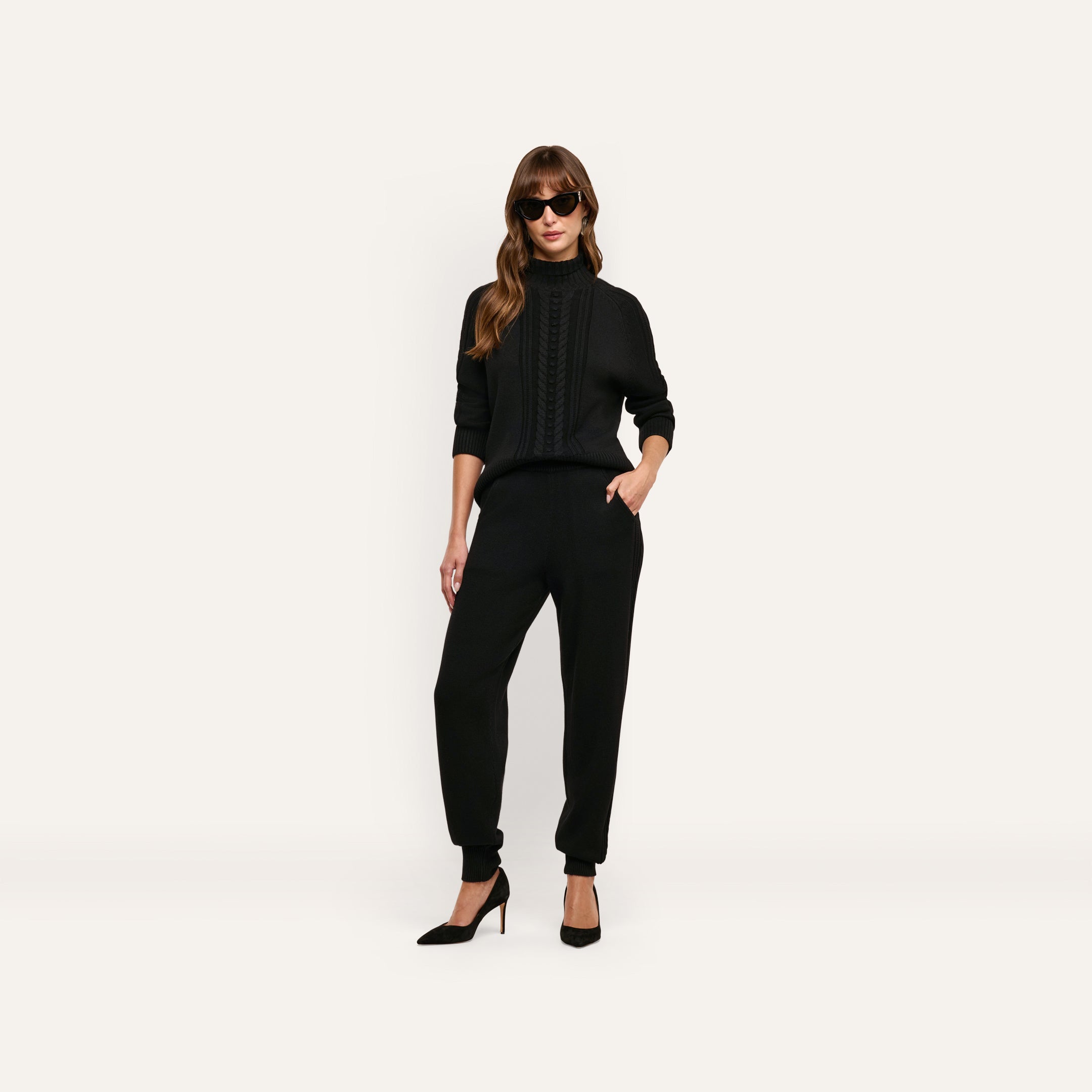 A woman wearing a black Sandrina Turtleneck cable-knit sweater, matching joggers, and black heels.