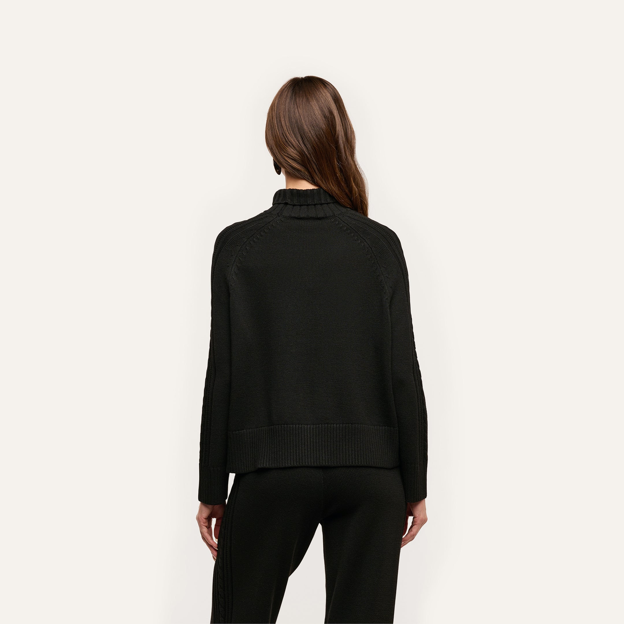 Back view of a person wearing a relaxed fit black cable knit Sandrina Turtleneck.