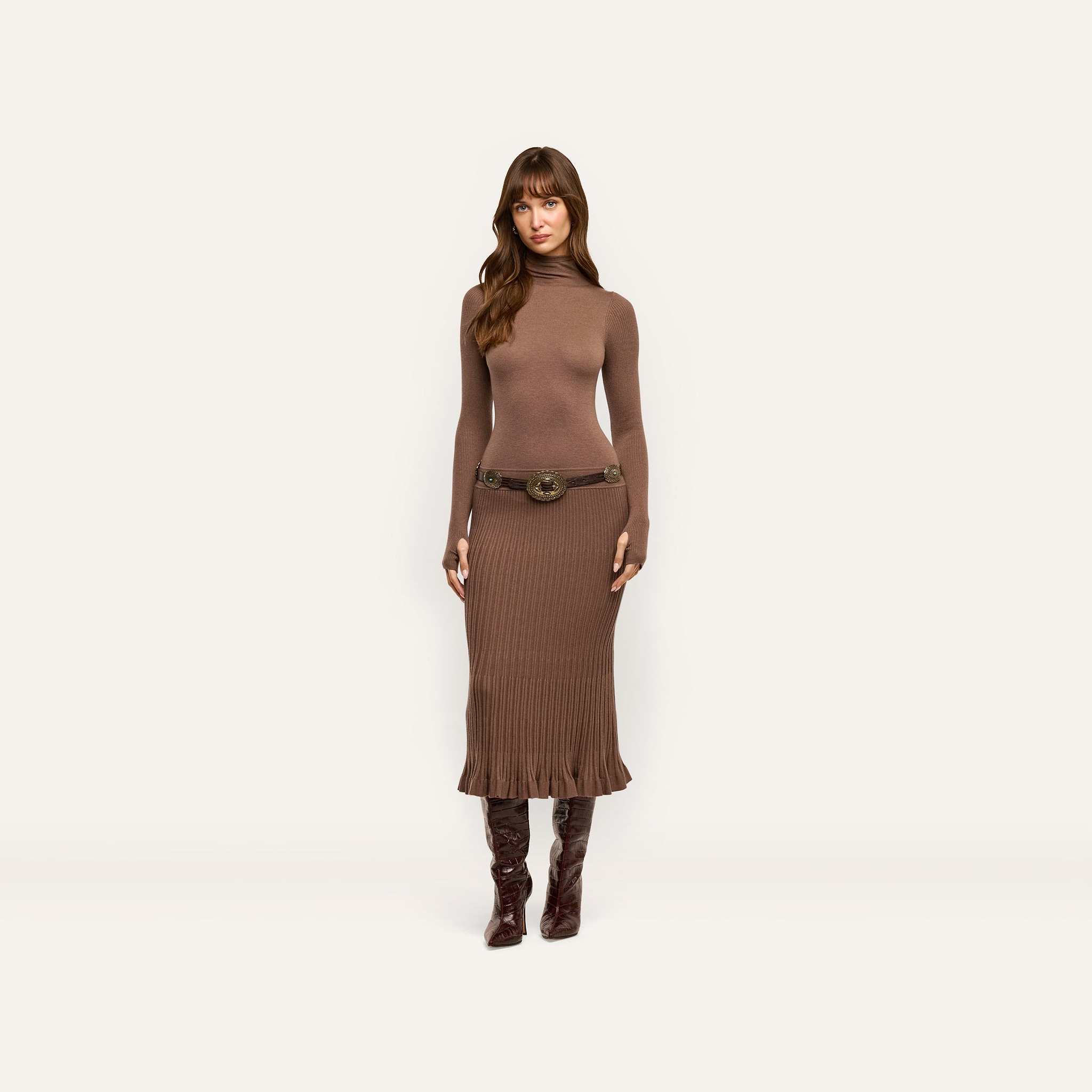 A person wearing a brown, lightweight Solaire Knit Dress featuring a funnelneck, long sleeves, a wide belt with an ornate buckle, and knee-high boots.