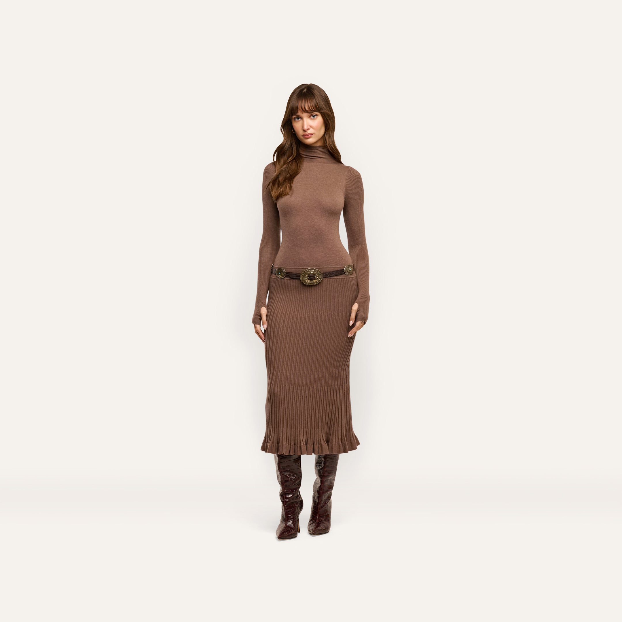 A person wearing a brown, lightweight Solaire Knit Dress featuring a funnelneck, long sleeves, a wide belt with an ornate buckle, and knee-high boots.