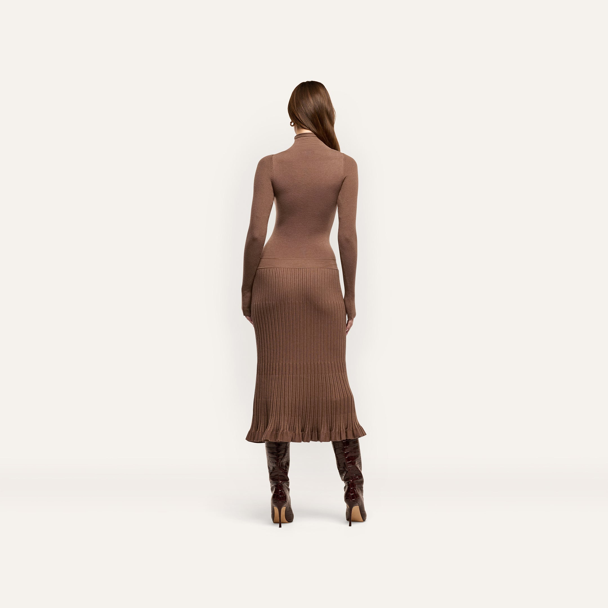 A person seen from behind, wearing a brown, long-sleeved, funnelneck Solaire Knit Dress in a lightweight midi knit paired with dark knee-high heeled boots.