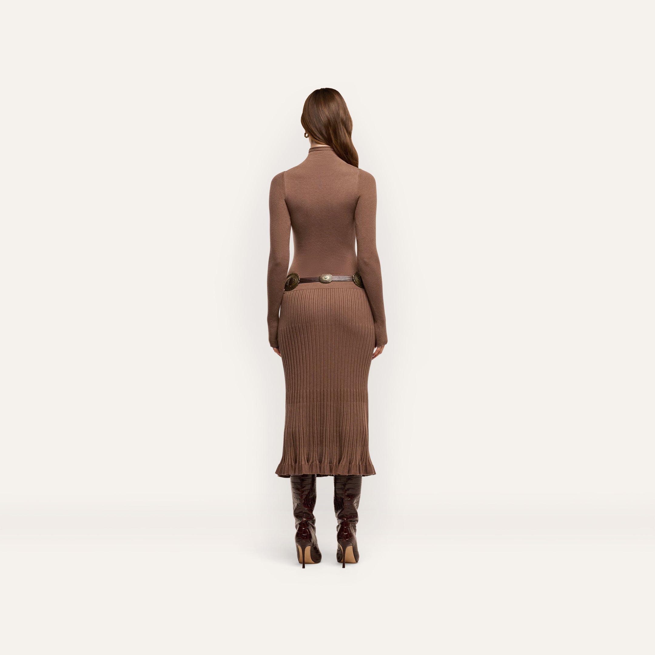 Back view of a person wearing a brown Solaire Knit Dress, a lightweight knit funnelneck midi design with a ribbed skirt, a thin metallic-buckle belt, and knee-high heeled boots.