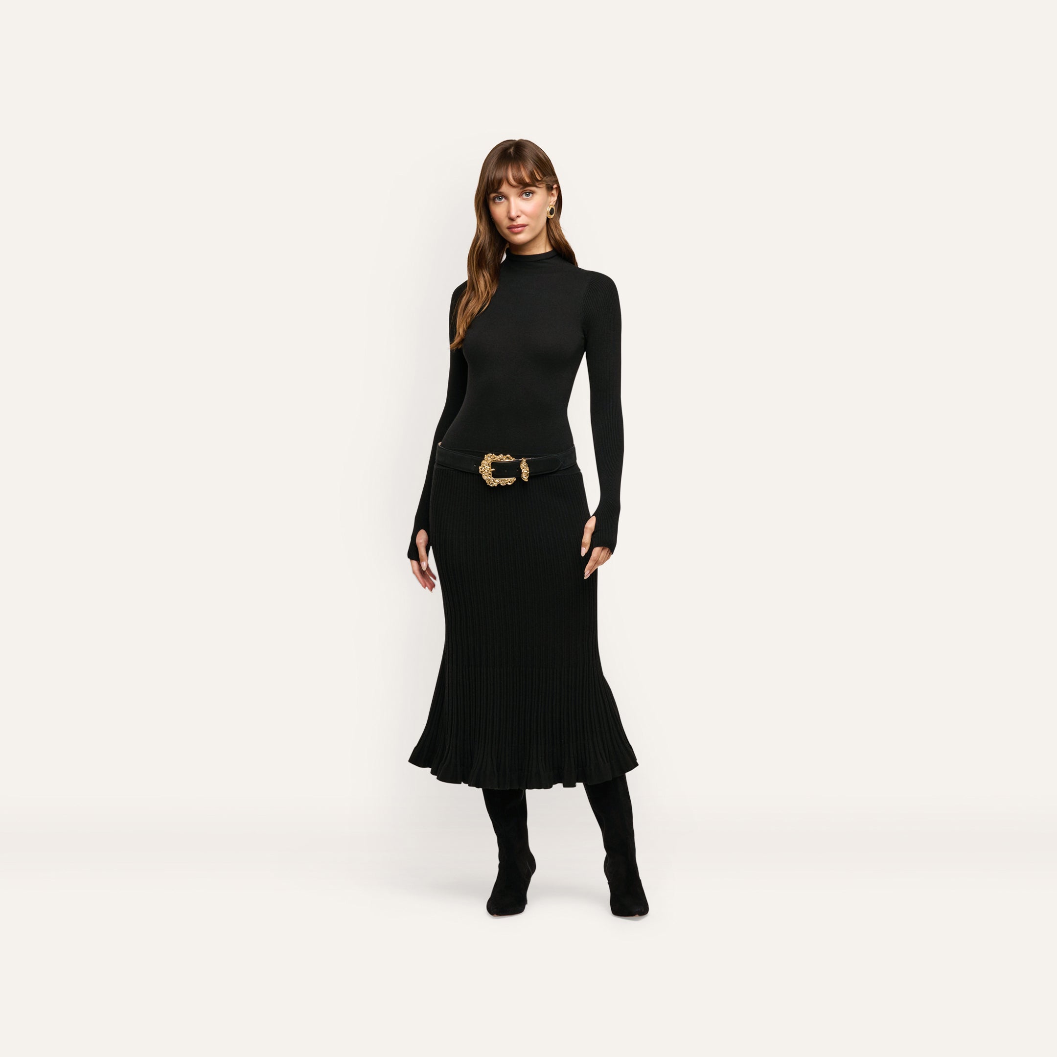 A person wears a black Solaire Knit Dress featuring a turtleneck, long sleeves with thumb holes, and a belt with a large gold ornate buckle.