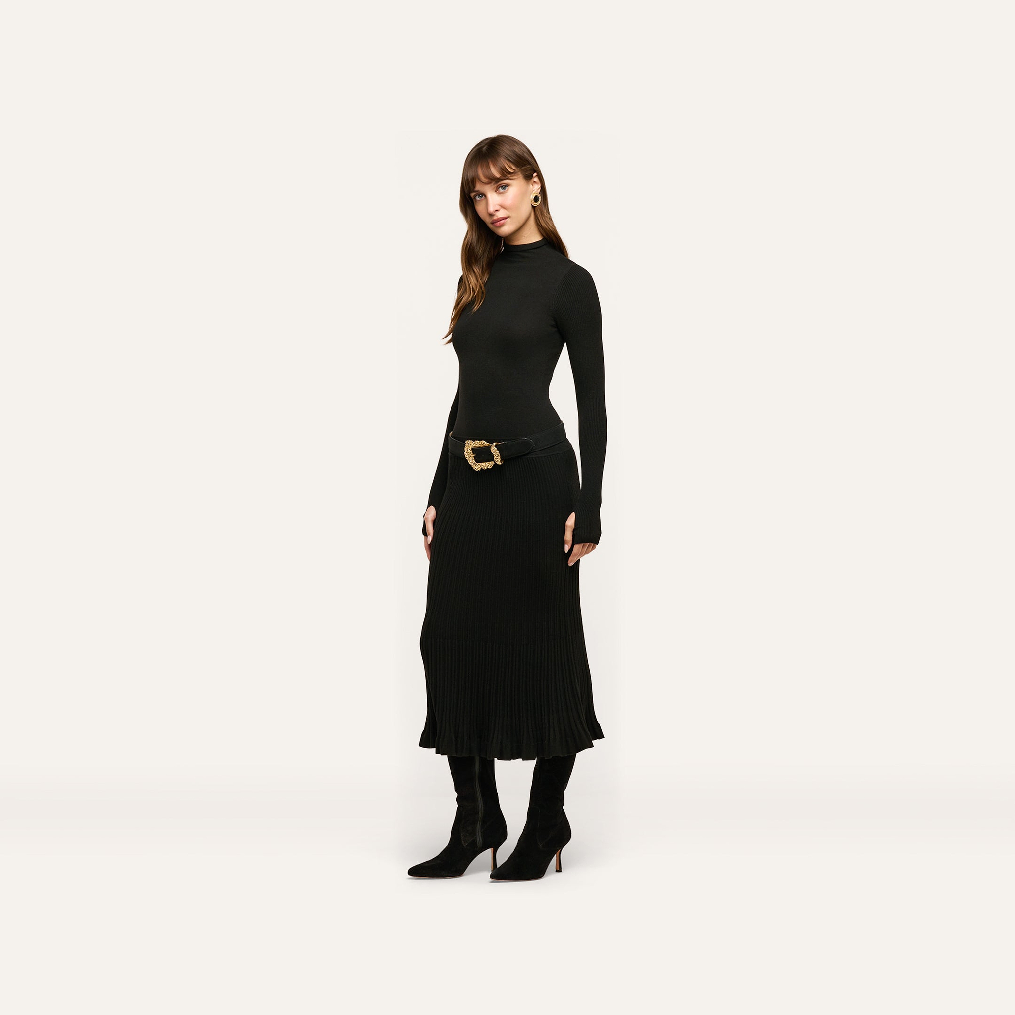 A woman wears the Solaire Knit Dress, a long-sleeved black midi dress in a lightweight knit, styled with a gold-buckled belt and black heeled boots.