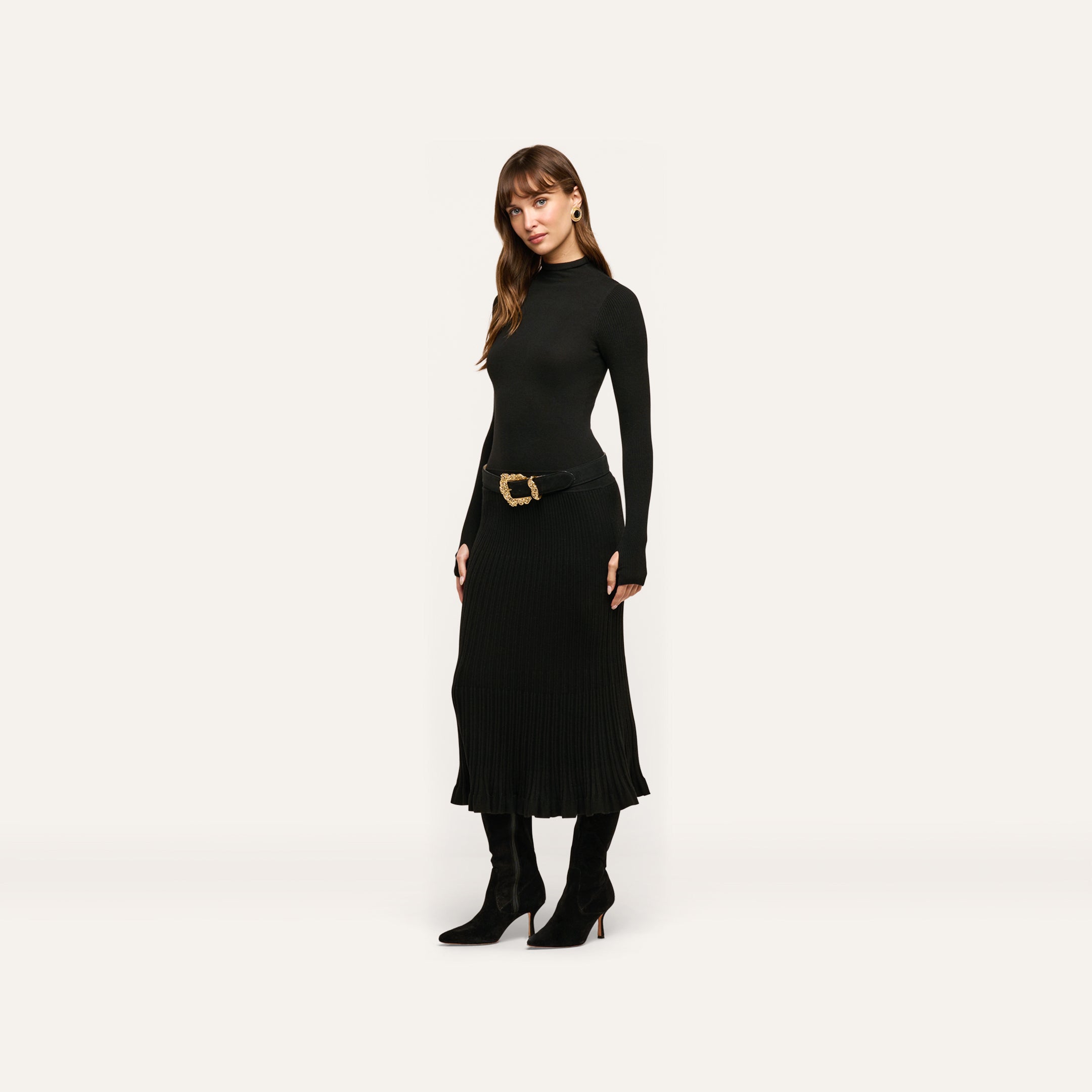 A woman wears the Solaire Knit Dress, a long-sleeved black midi dress in a lightweight knit, styled with a gold-buckled belt and black heeled boots.