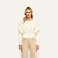 A woman wearing the cream-colored Ottavia Pullover, featuring a cable knit pattern and fringe trim on the shoulders, paired with tan pants.