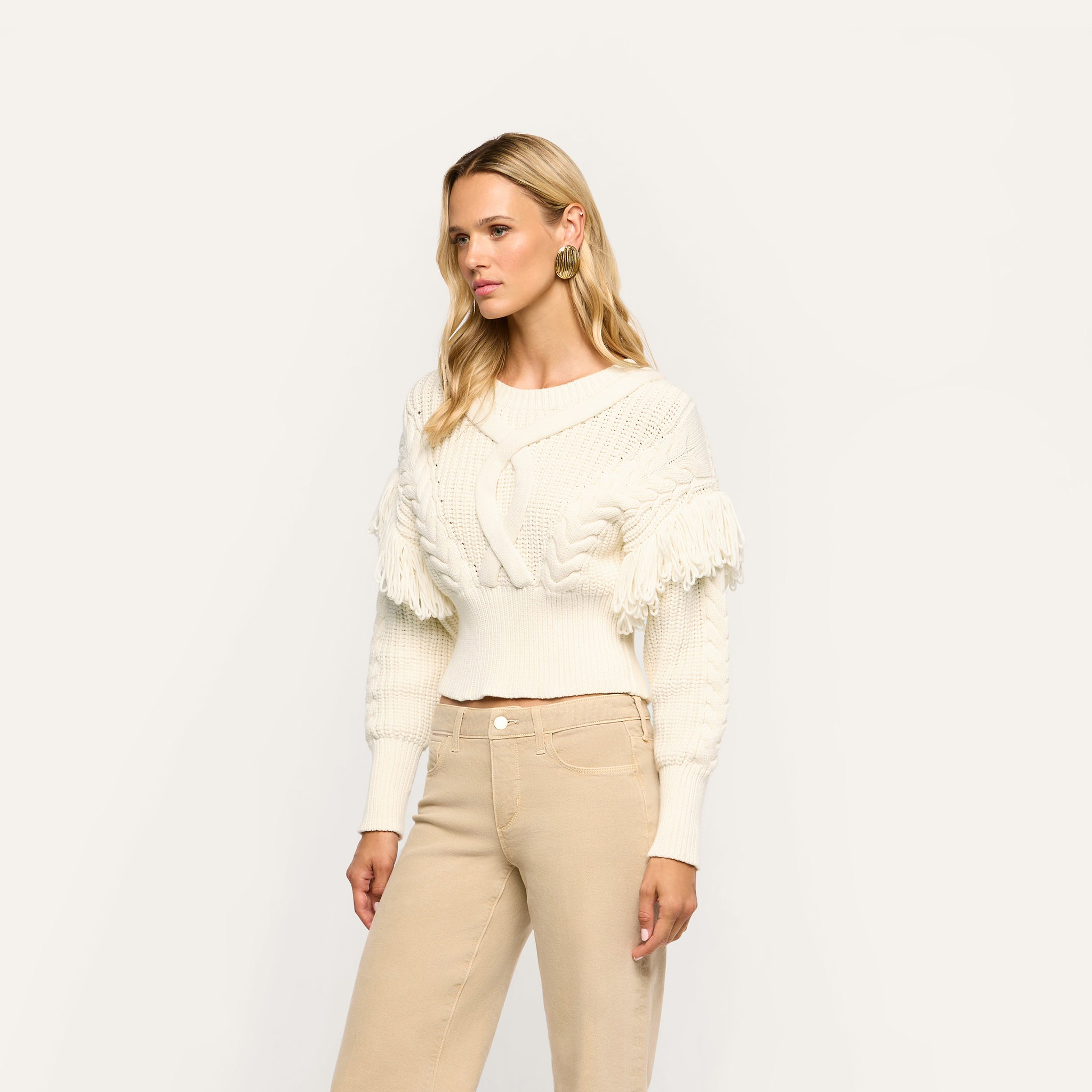 Cream-colored Ottavia Pullover featuring cable knit texture and fringe trim on the shoulders, worn with tan pants.