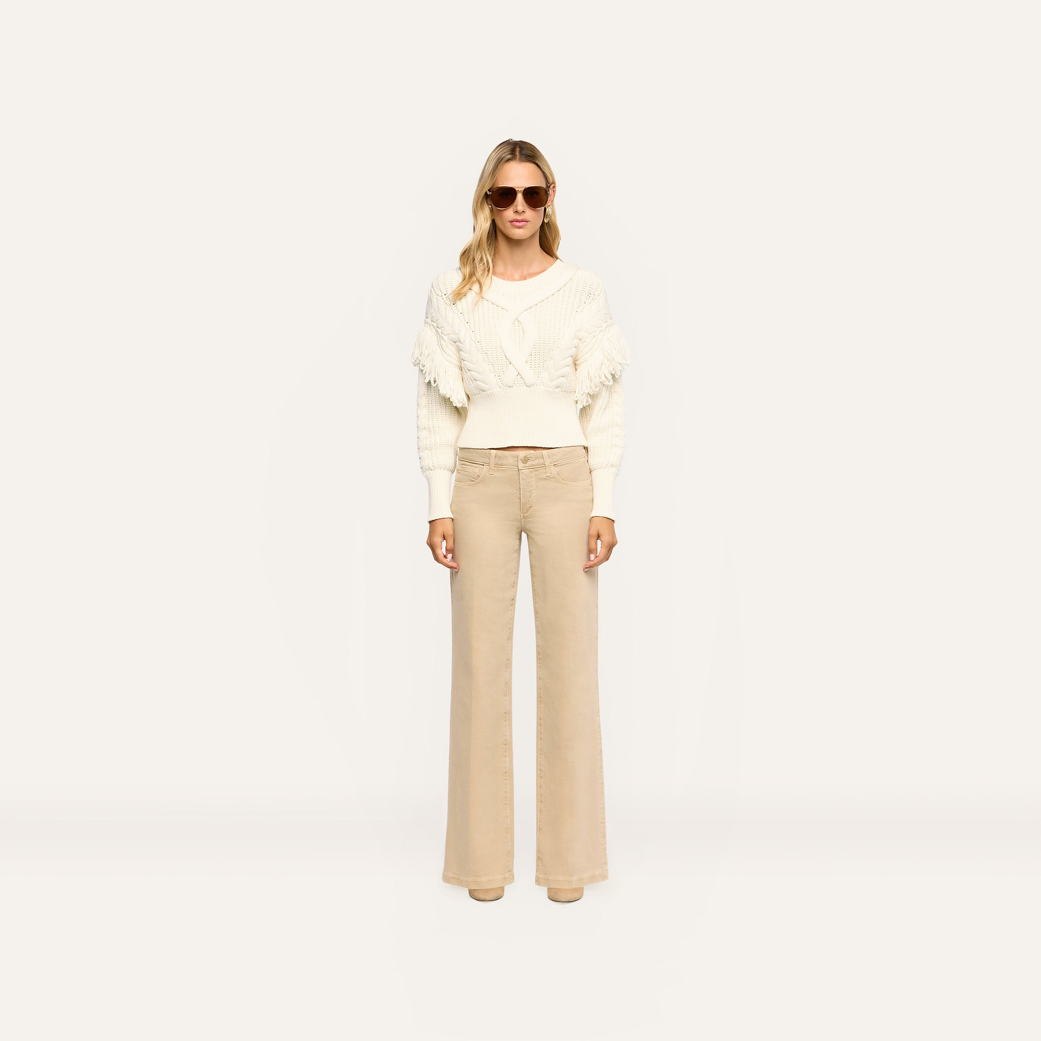 Woman wearing an Ottavia Pullover in cream-colored cable knit with fringe trim on the sleeves, paired with wide-leg tan trousers.