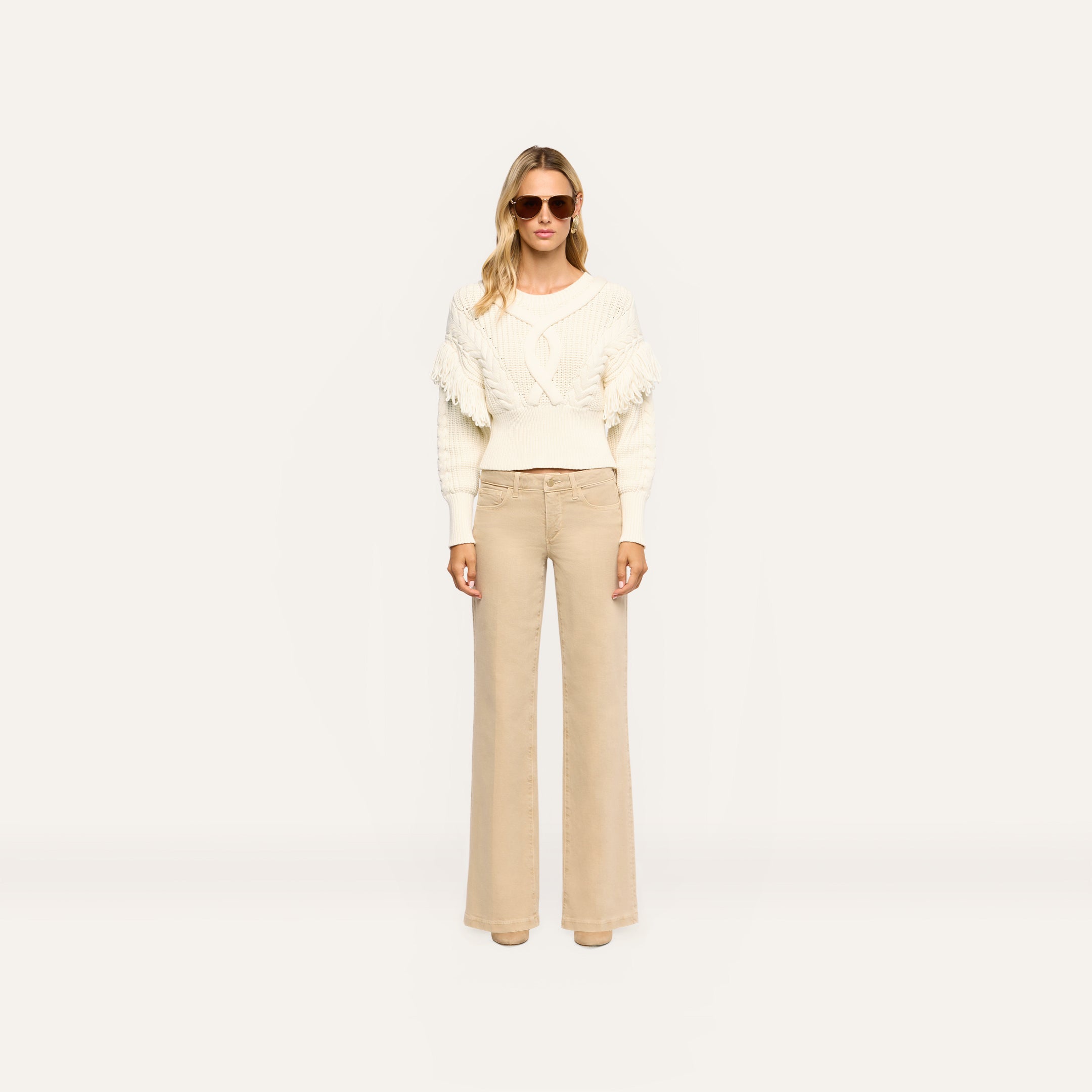 Woman wearing an Ottavia Pullover in cream-colored cable knit with fringe trim on the sleeves, paired with wide-leg tan trousers.