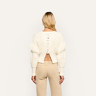 Back view of the Ottavia Pullover, a cream-colored cable knit sweater with a lace-up back cutout, fringe trim on the shoulders, and tan pants.
