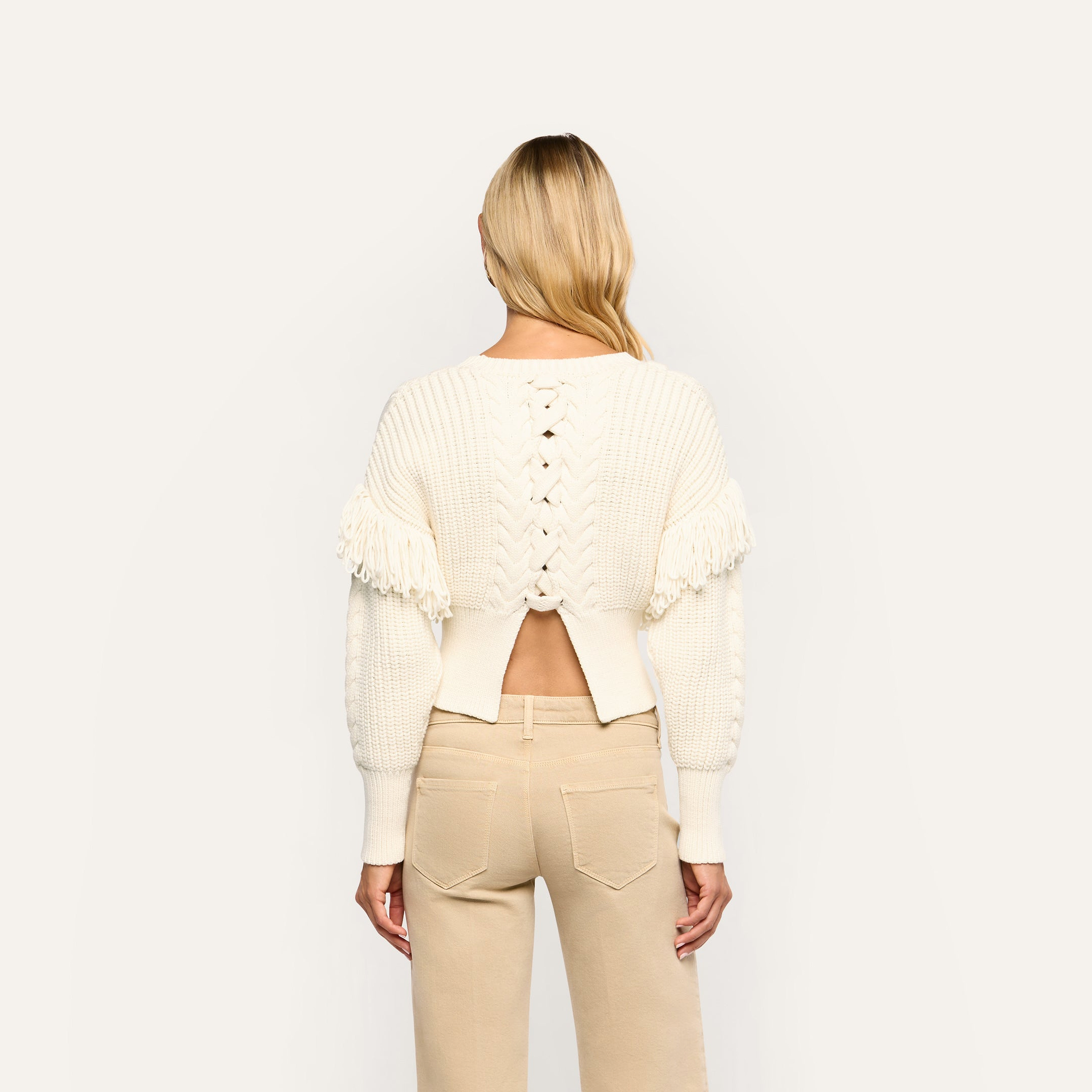 Back view of the Ottavia Pullover, a cream-colored cable knit sweater with a lace-up back cutout, fringe trim on the shoulders, and tan pants.