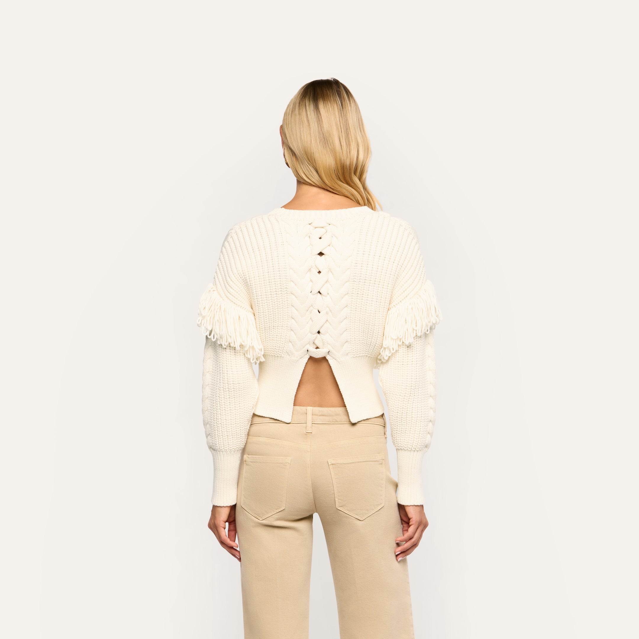 Back view of the Ottavia Pullover, a cream-colored cable knit sweater with a lace-up back cutout, fringe trim on the shoulders, and tan pants.
