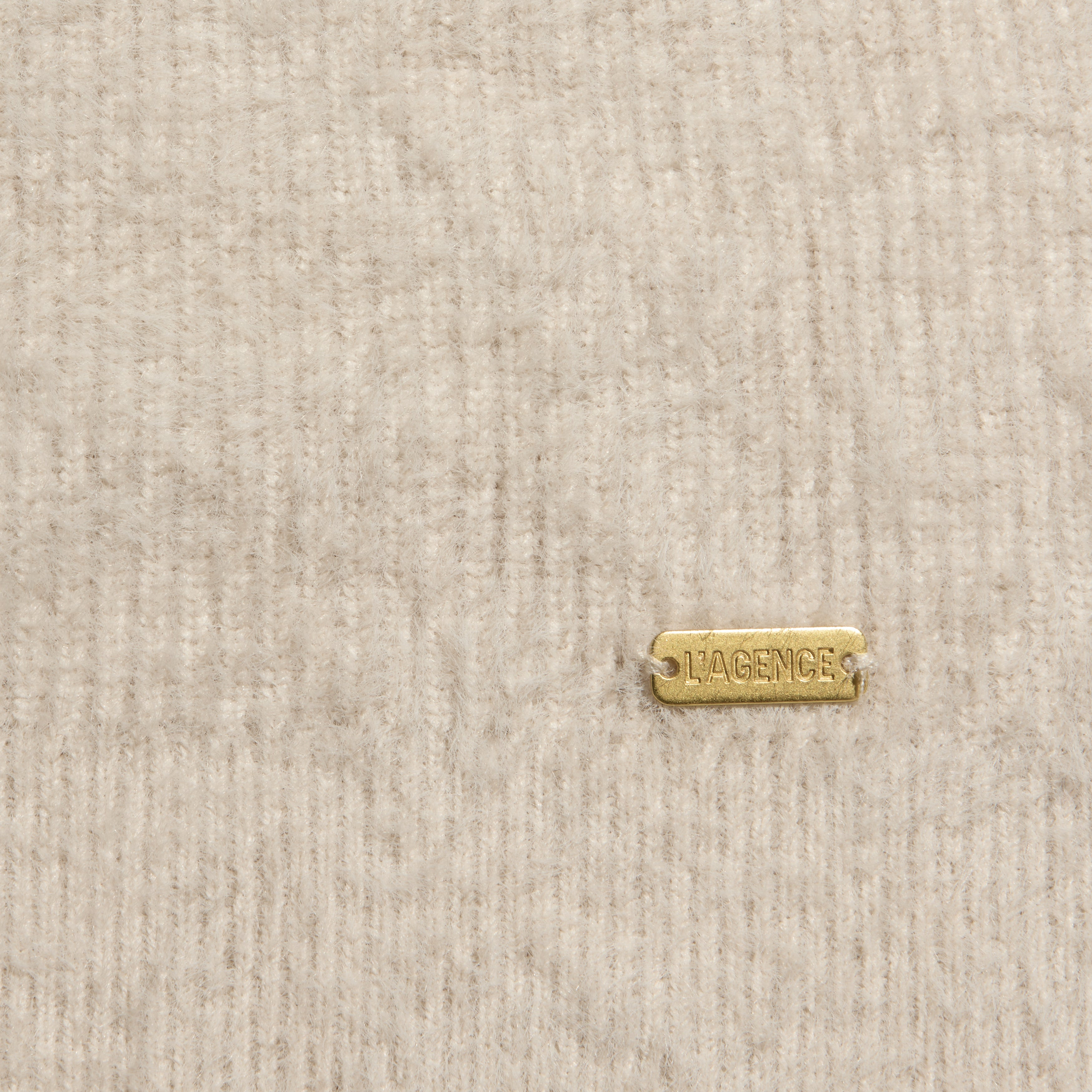 Close-up of a fuzzy, ribbed beige Korinna Pullover featuring a small rectangular gold-tone metal tag engraved with LAGENCE.